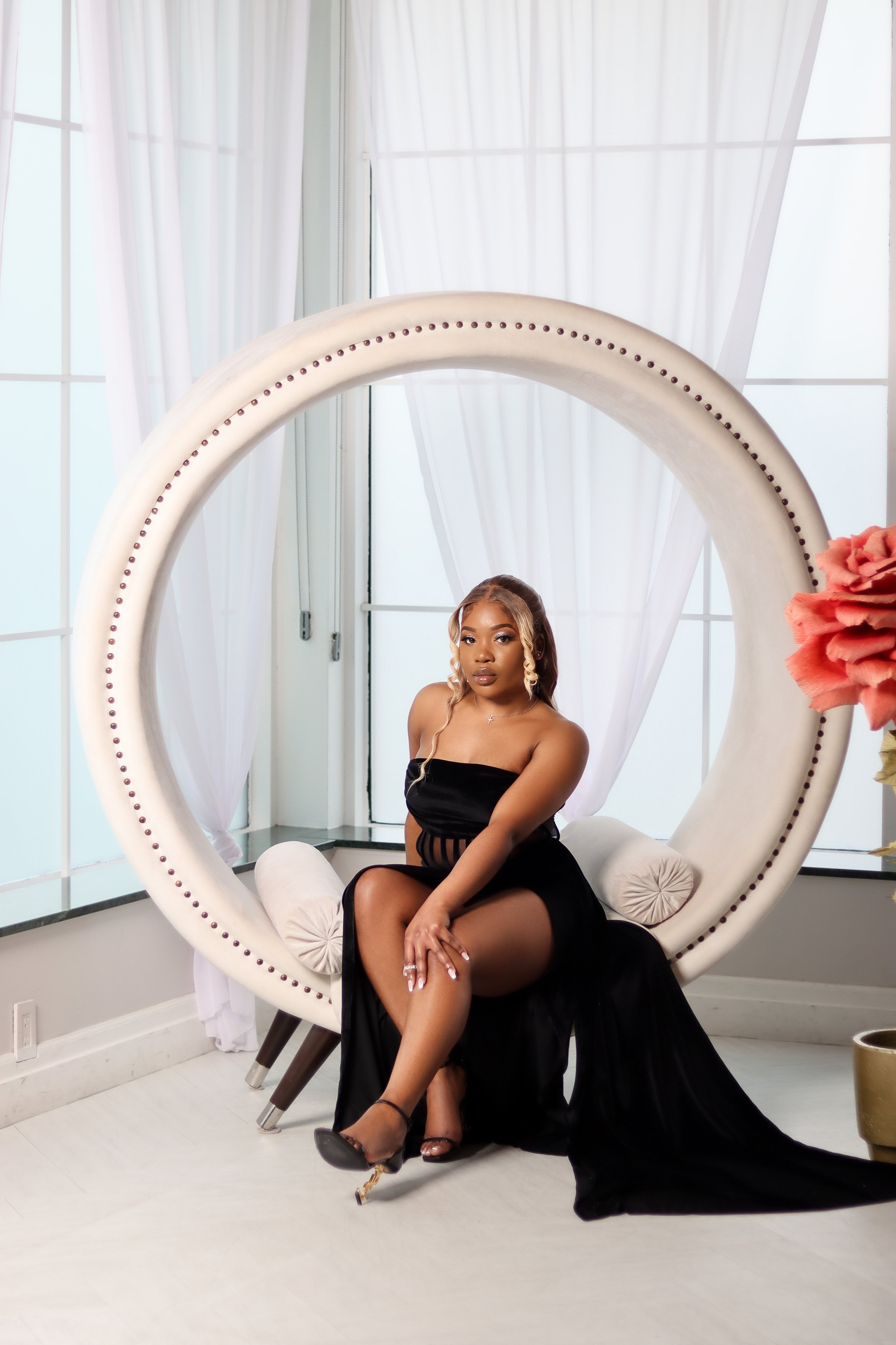A woman in a black dress sitting on a white, circular, modern chair with a large semi-circular back and small vertical embellishments, in front of large windows with white curtains, in a bright, elegant room with a light floor, and pink flowers on th