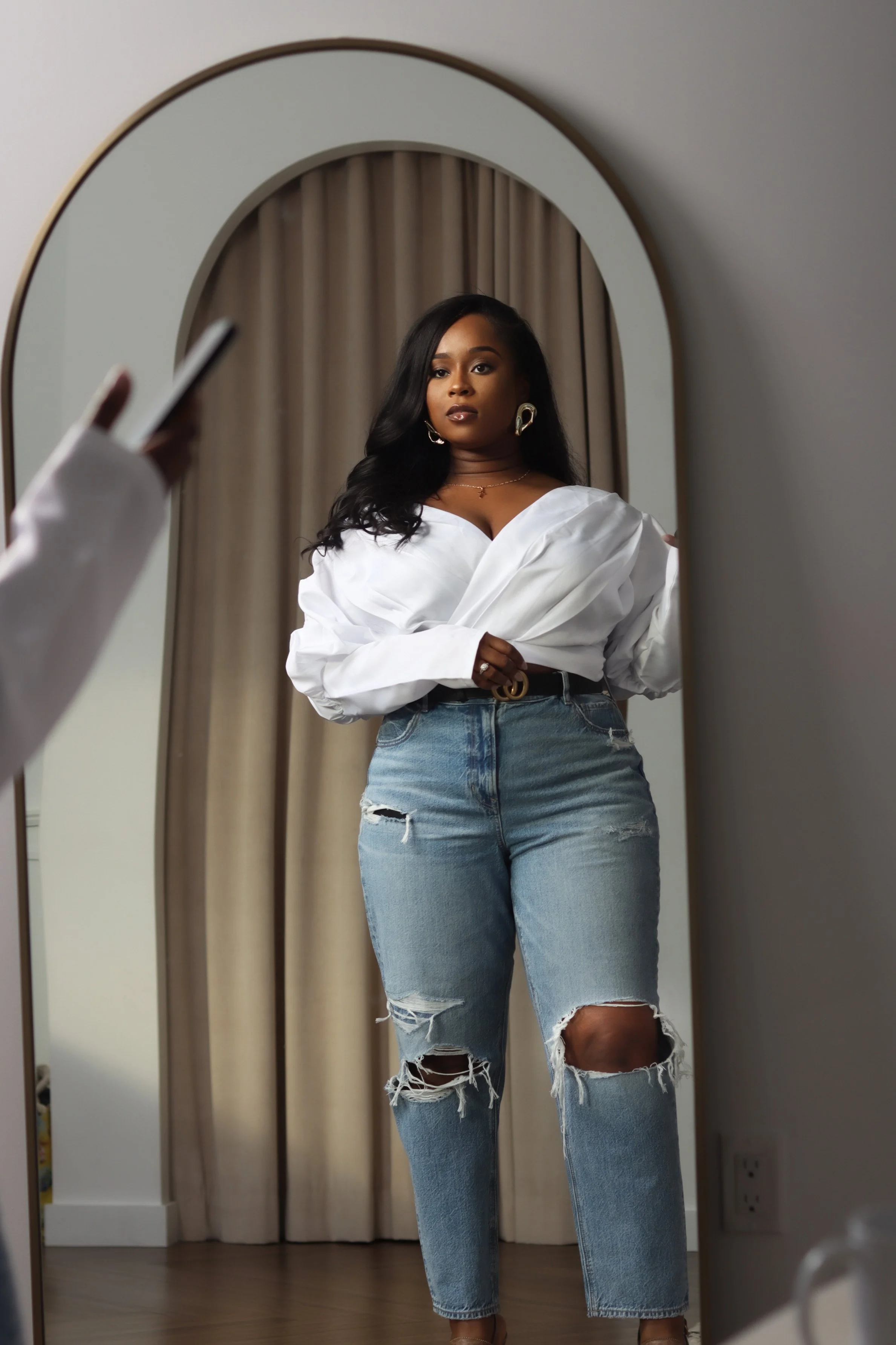 A woman taking a photo of herself in a mirror, wearing a white top and ripped jeans with large rips at the knees.