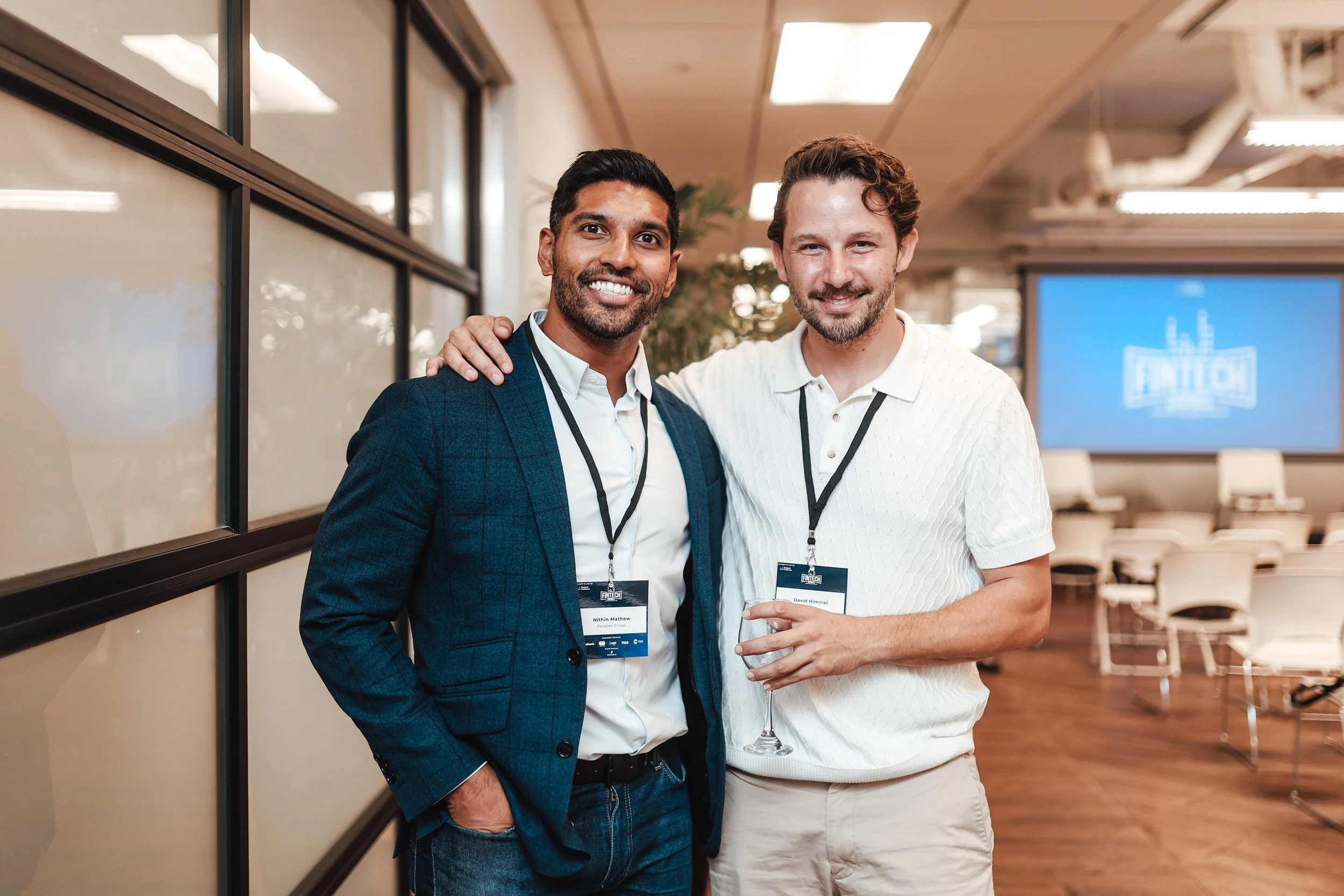 Two men smiling at a conference, one with a beard and dark hair wearing a dark suit, the other with light brown hair in a white shirt holding a glass, standing in a conference room with chairs and a large screen in the background.