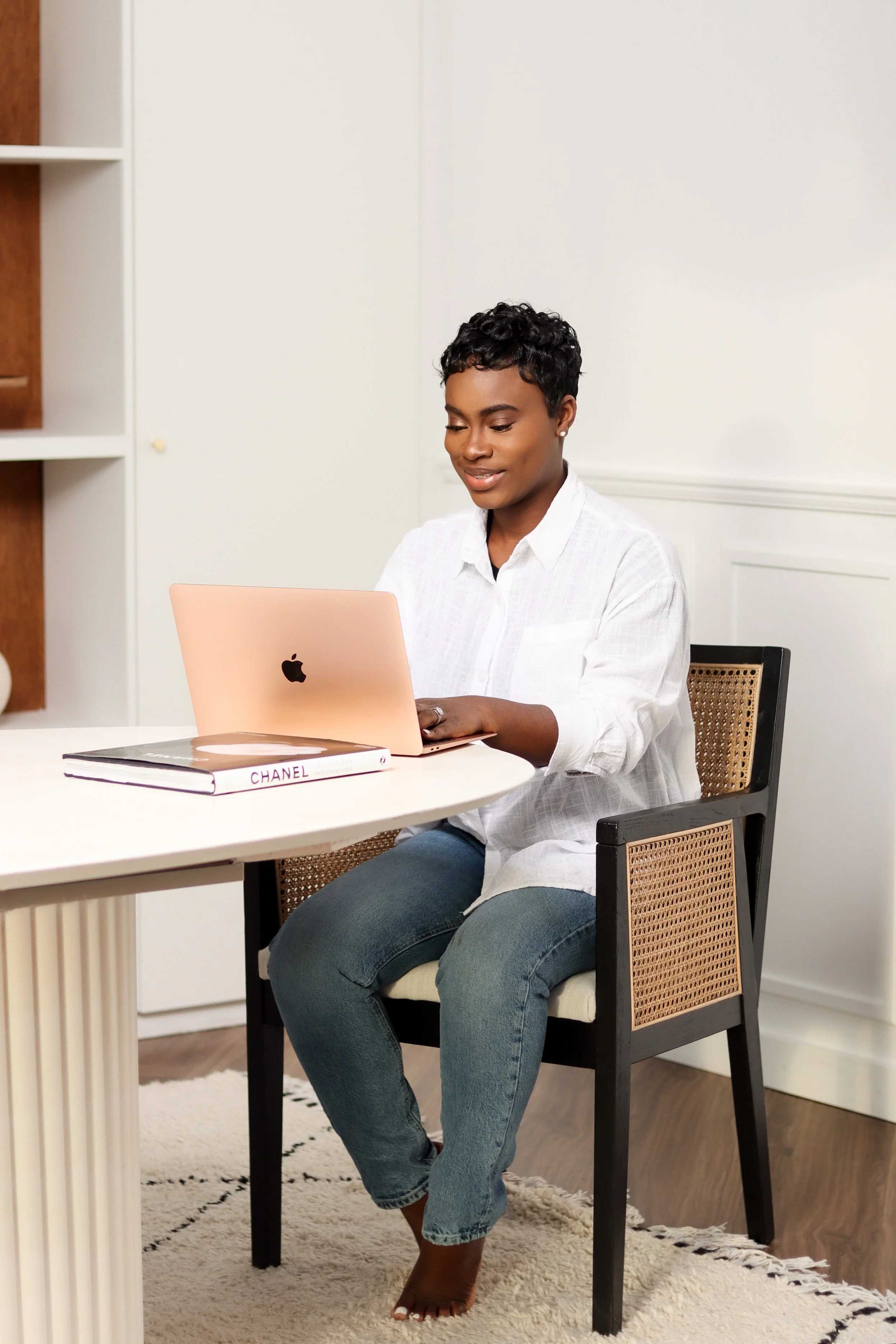 Woman sitting at a desk using a pink Apple MacBook with a Chanel notebook nearby, in a modern, minimalistic room.