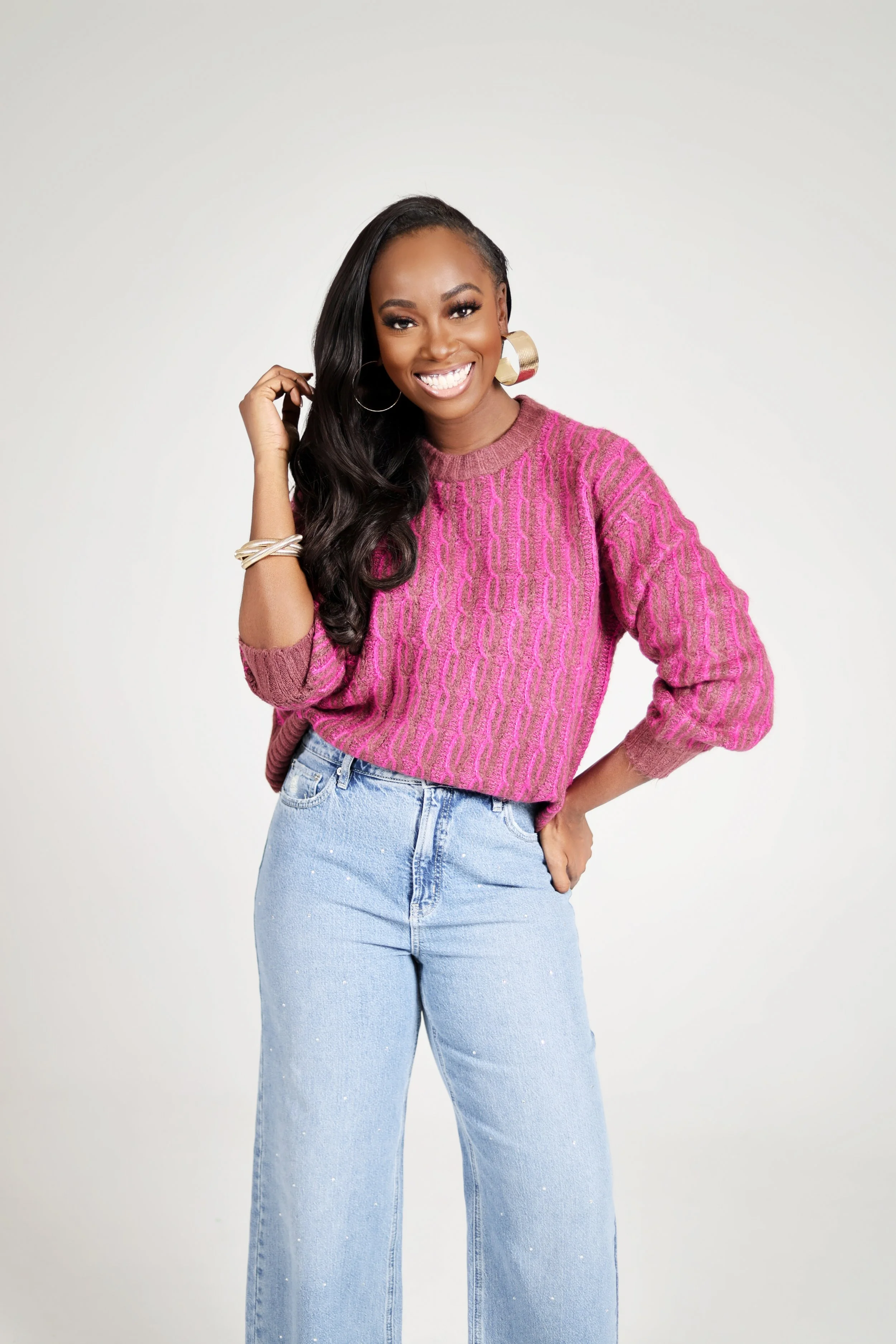 A smiling woman with long black hair, wearing a pink sweater and light blue jeans, standing against a plain white background.