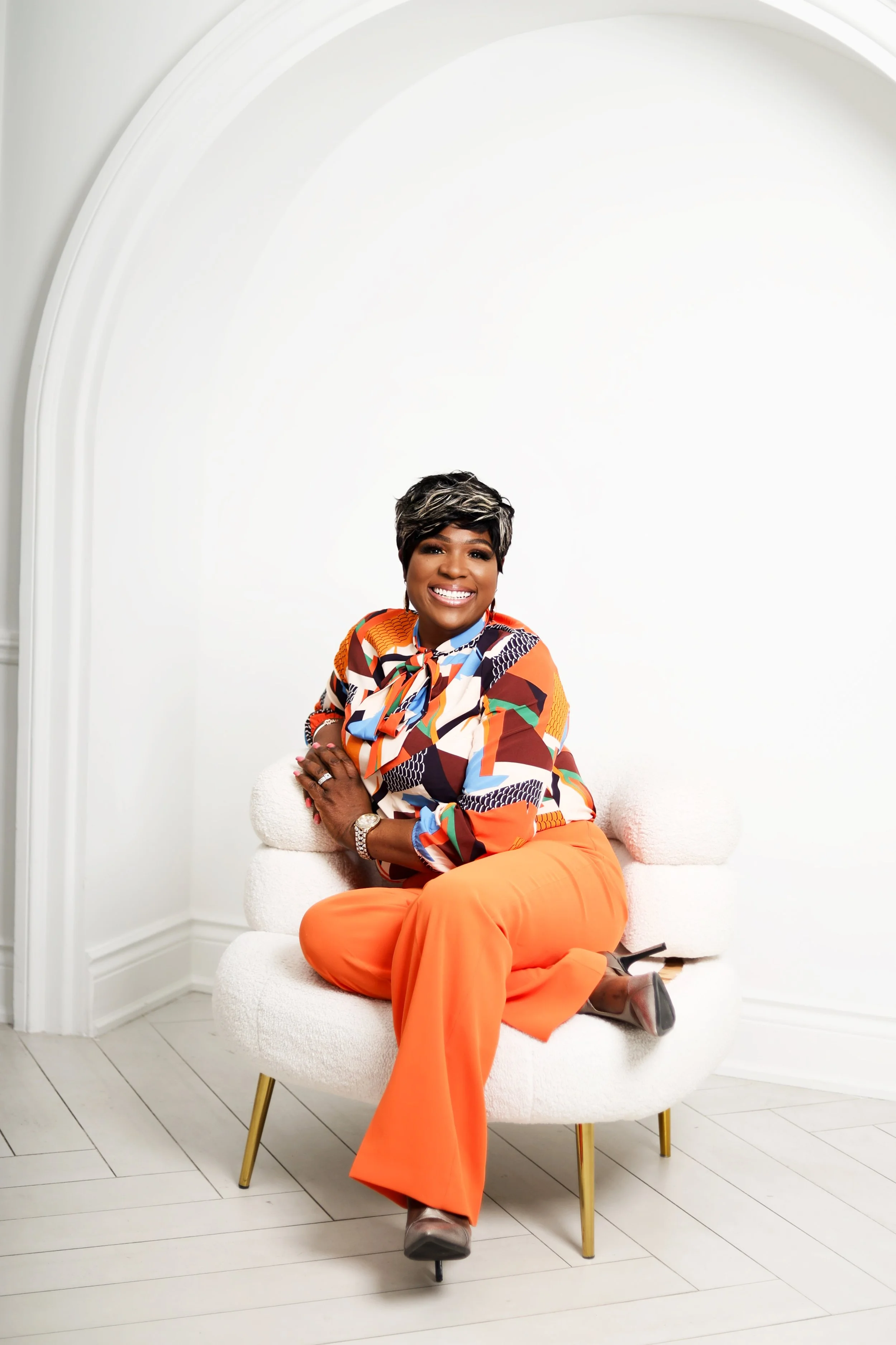 A smiling woman with short hair in a stylish colorful blouse and orange pants, sitting cross-legged on a modern white chair with gold legs in a bright room with white walls and flooring.