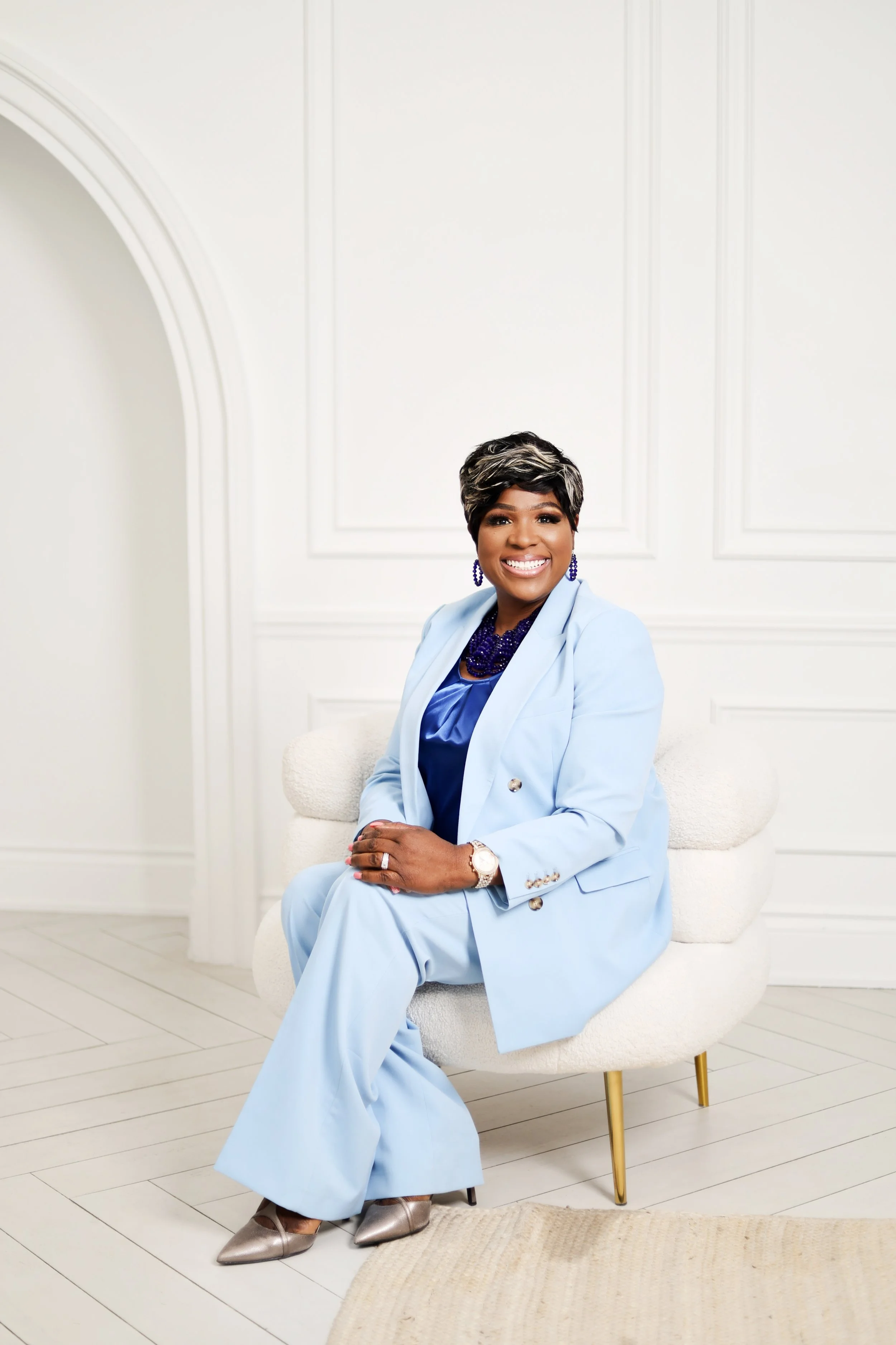 Woman in a light blue suit sitting on a white chair in a bright room, smiling at the camera.