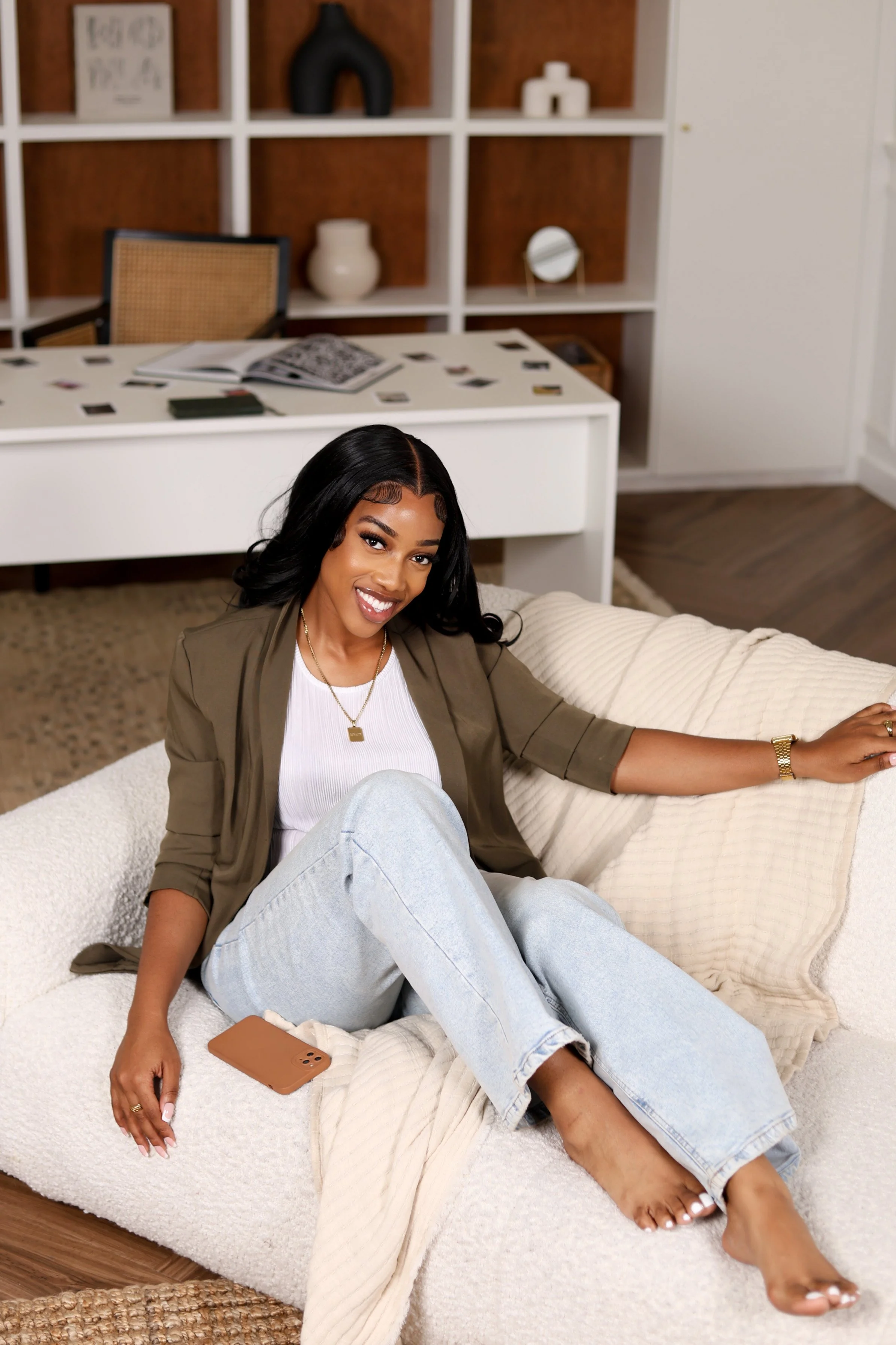 A woman sitting comfortably on a light-colored sofa with a beige pillow, smiling at the camera, in a modern living room with a white desk and decorative shelves in the background.