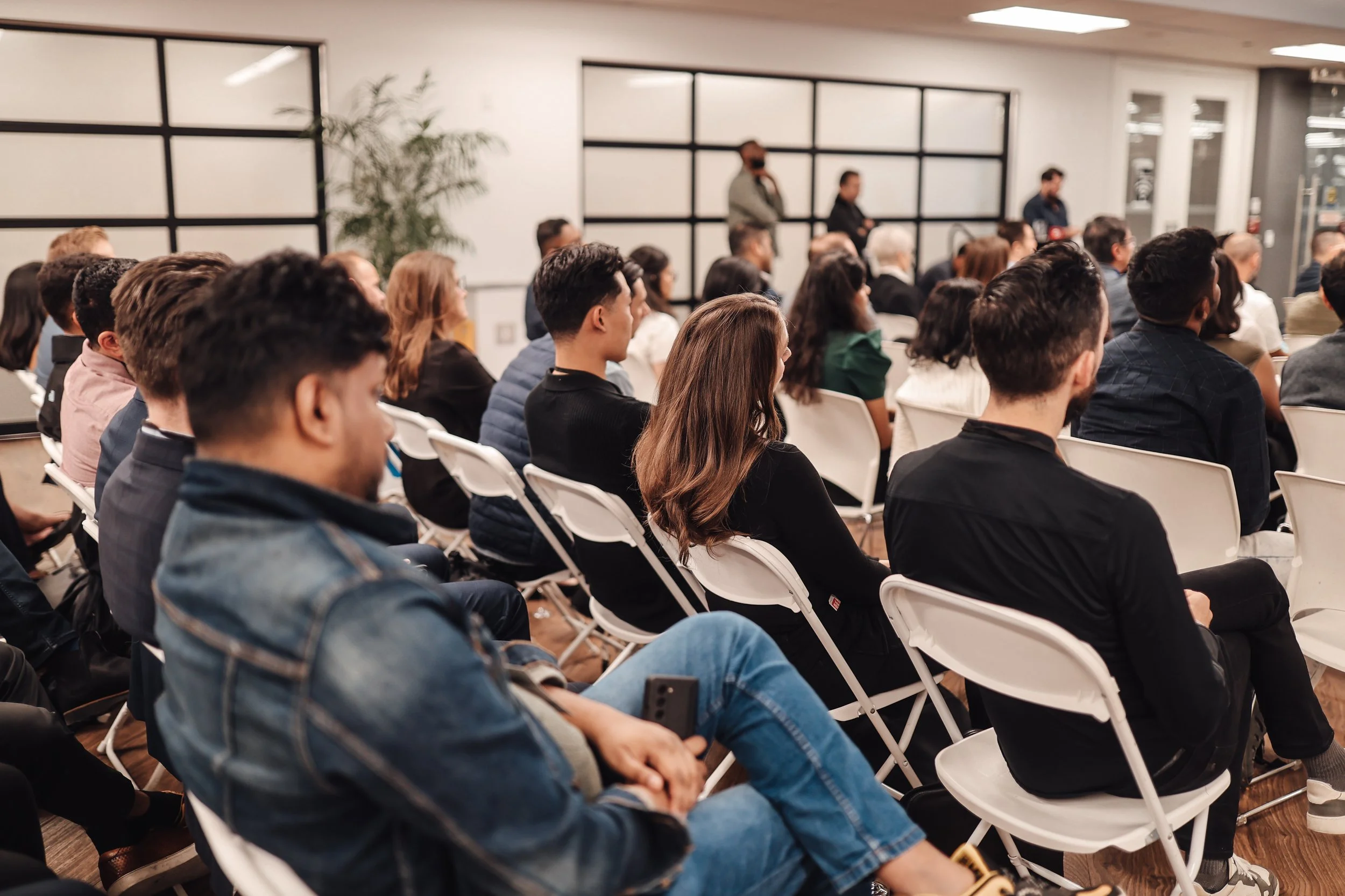 People attending a conference or seminar in a modern indoor venue, seated in white chairs, facing a speaker or presentation at the front.