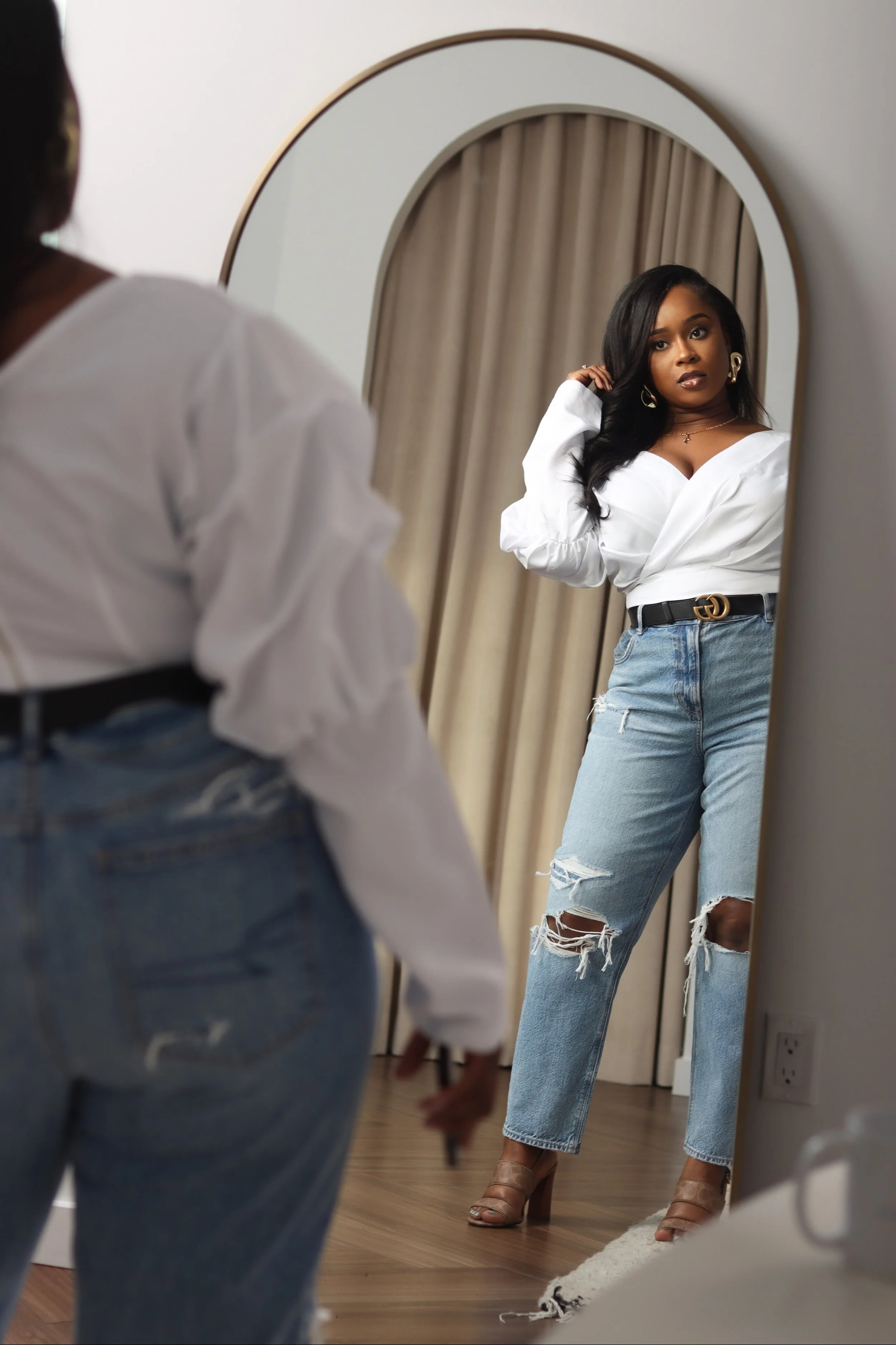 A woman looking at herself in a large full-length mirror, wearing a white shirt, ripped jeans, and high heels, with curtains in the background.