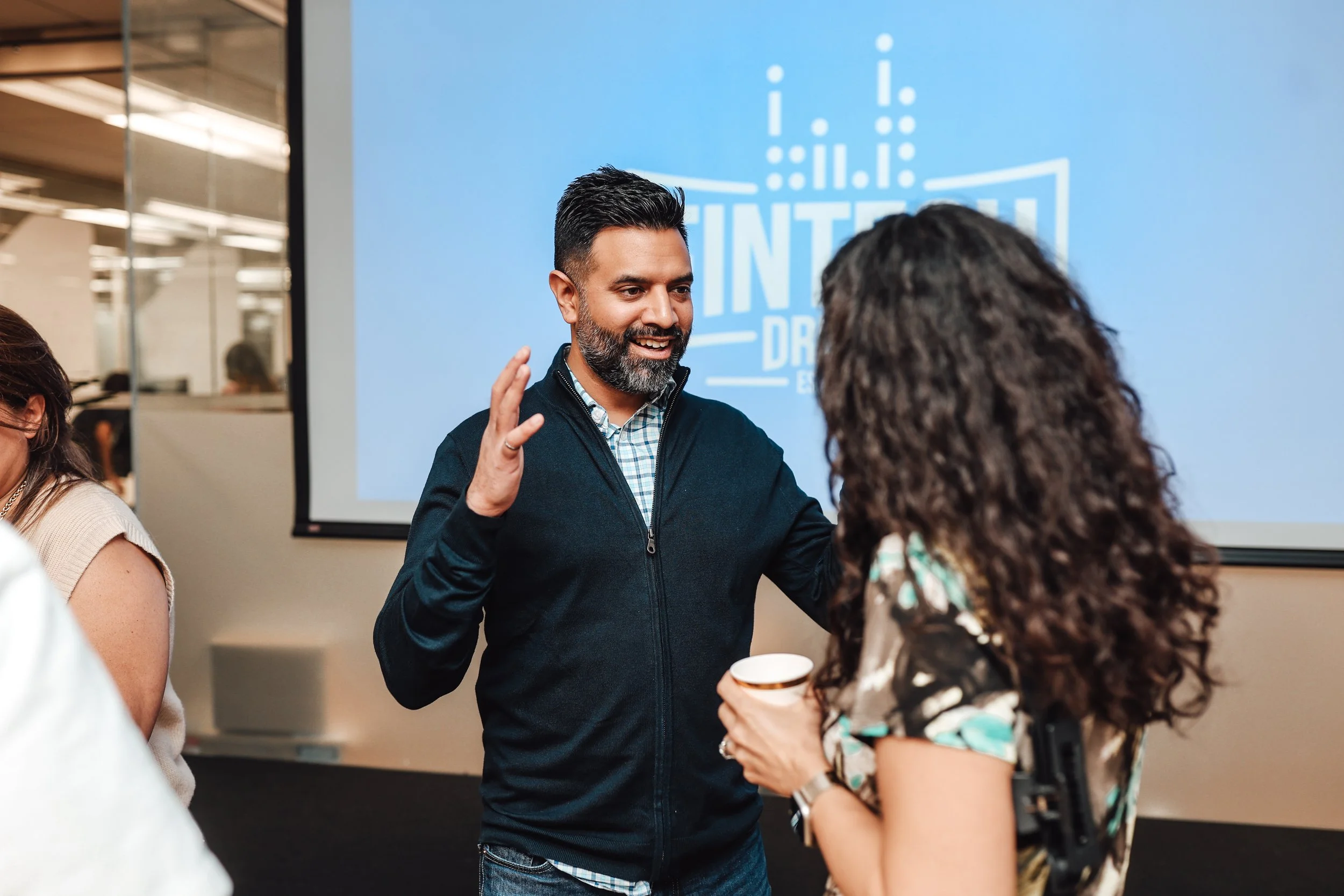 A man with a beard and dark hair, wearing a dark jacket and checkered shirt, is talking to a woman with curly brown hair, holding a paper cup, at a social event or conference with a blue presentation screen in the background.
