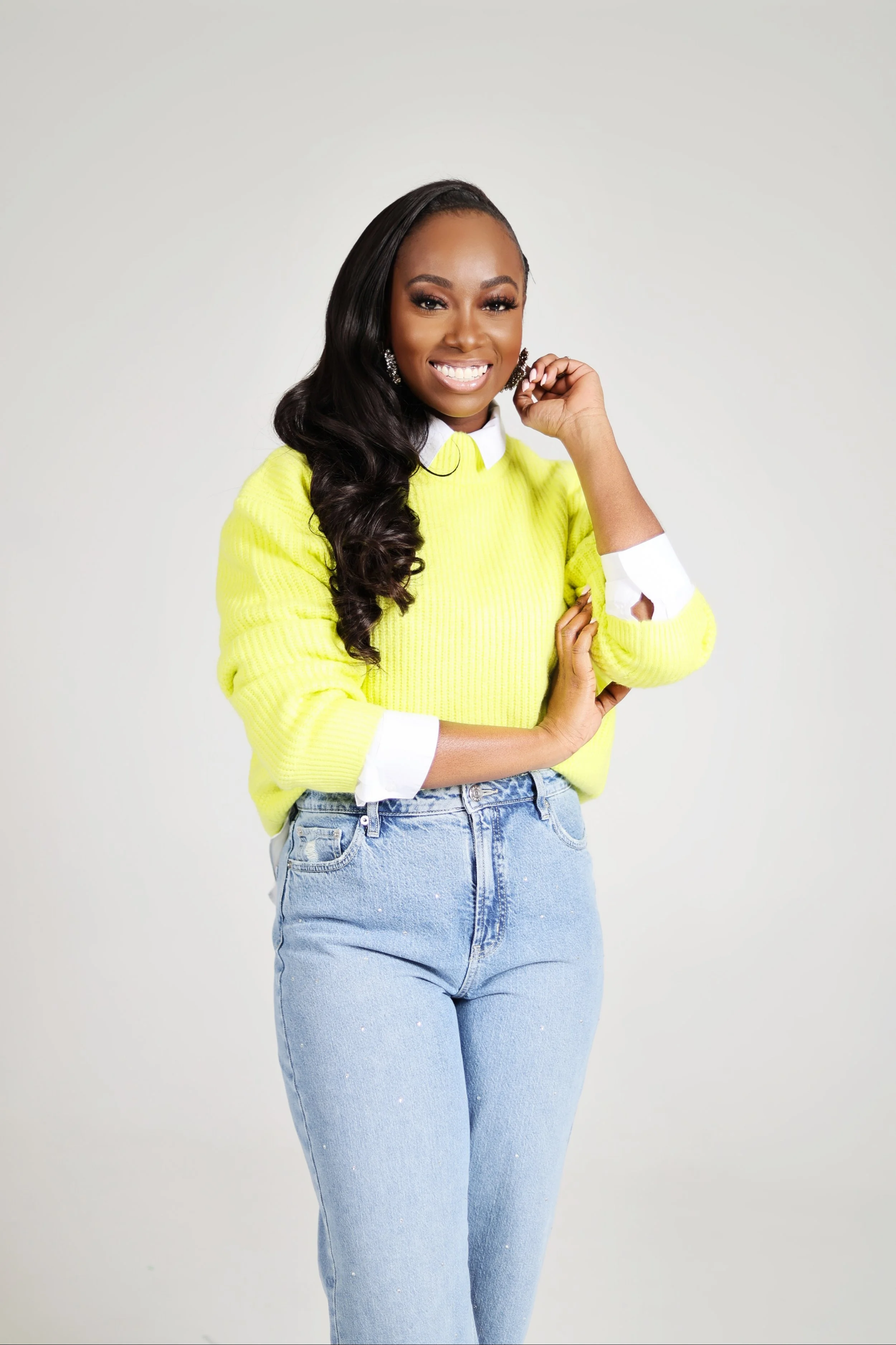 Portrait of a smiling woman with long dark hair, wearing a yellow sweater, white collared shirt underneath, and light blue jeans, standing against a plain light background.