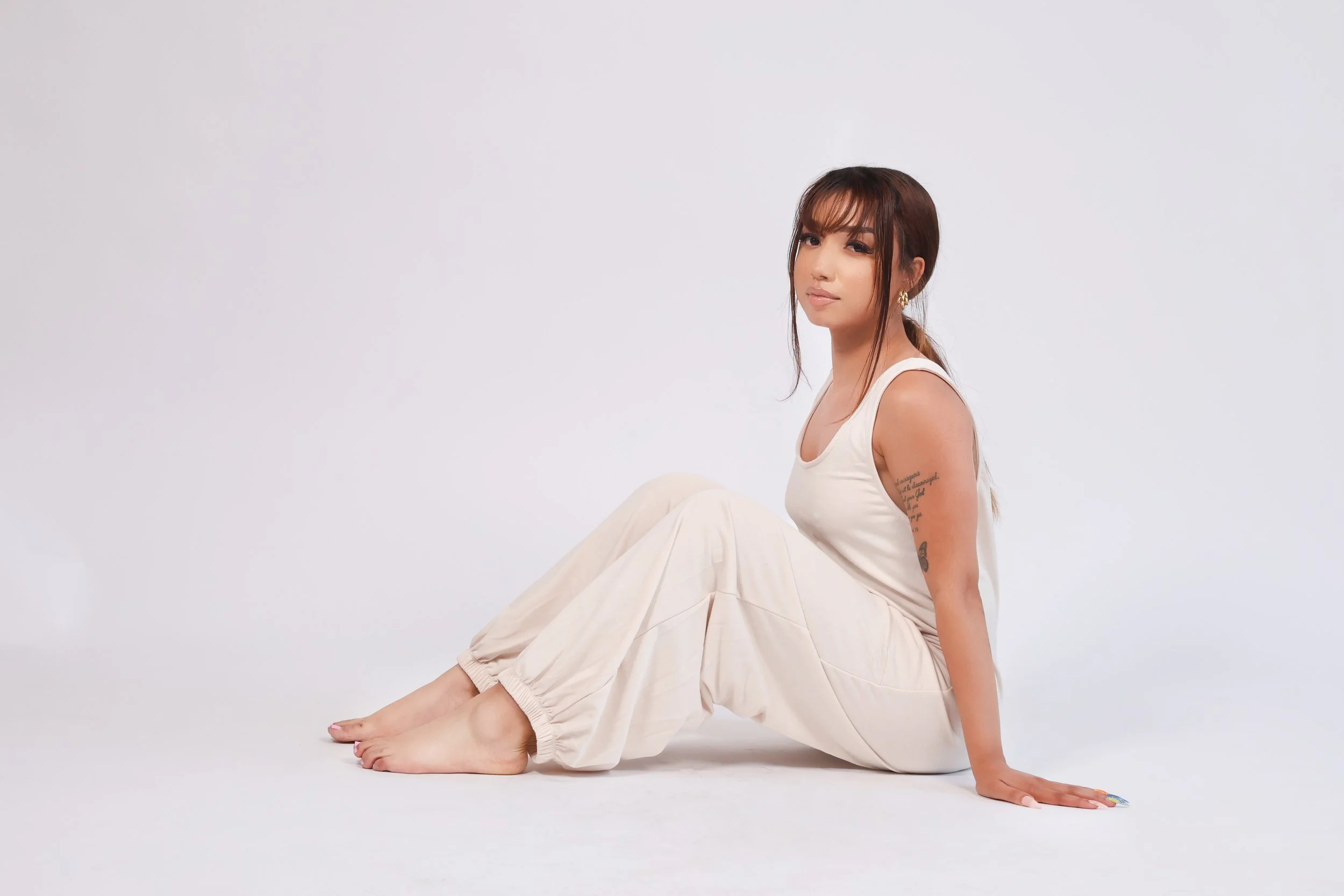 A woman sitting on the floor against a white background, wearing a white tank top and beige pants, with tattoos on her arm.