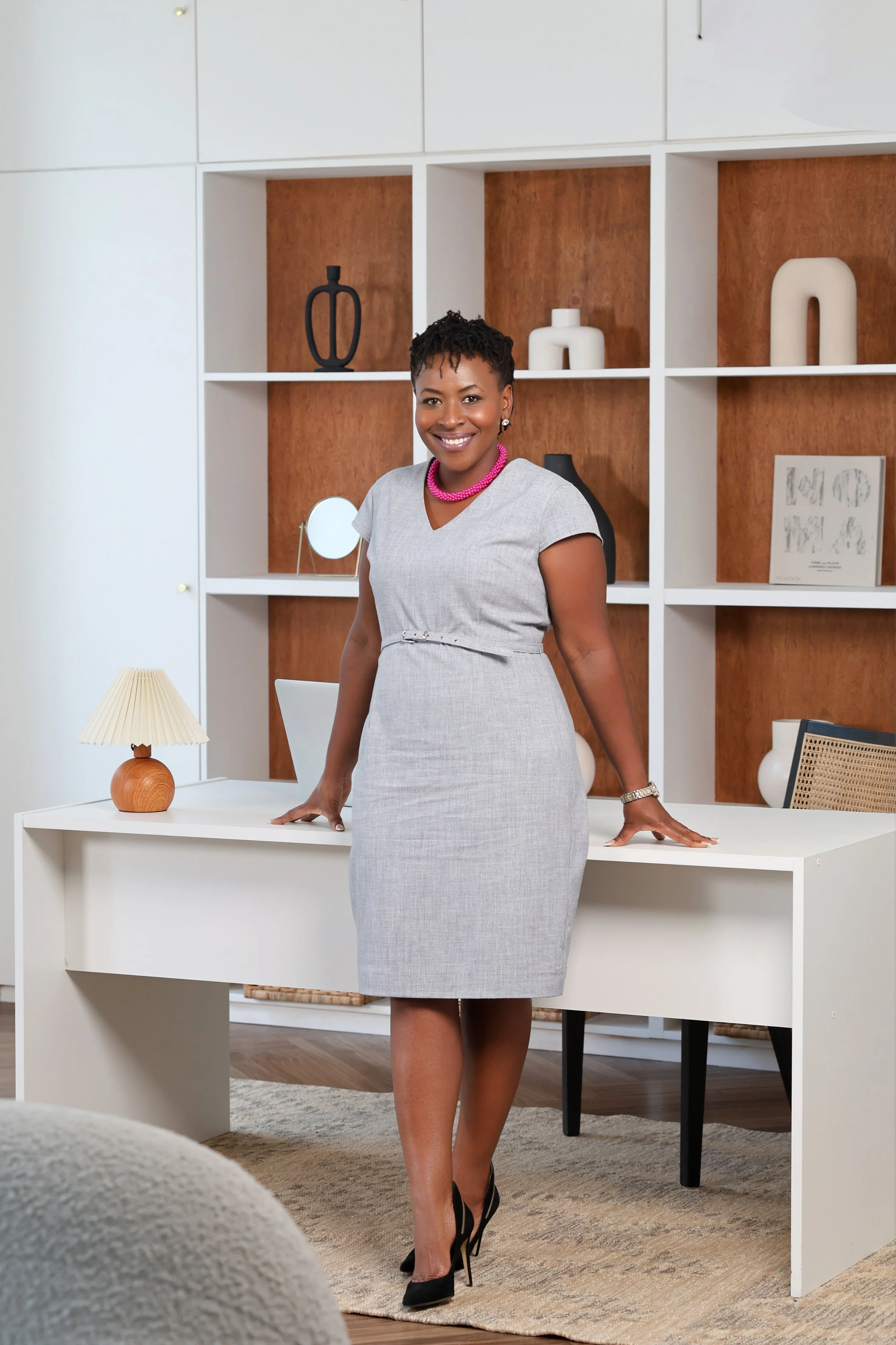 A woman in a gray dress with black high heels and a pink necklace is standing behind a white desk in a modern office space with wooden and white shelving units.