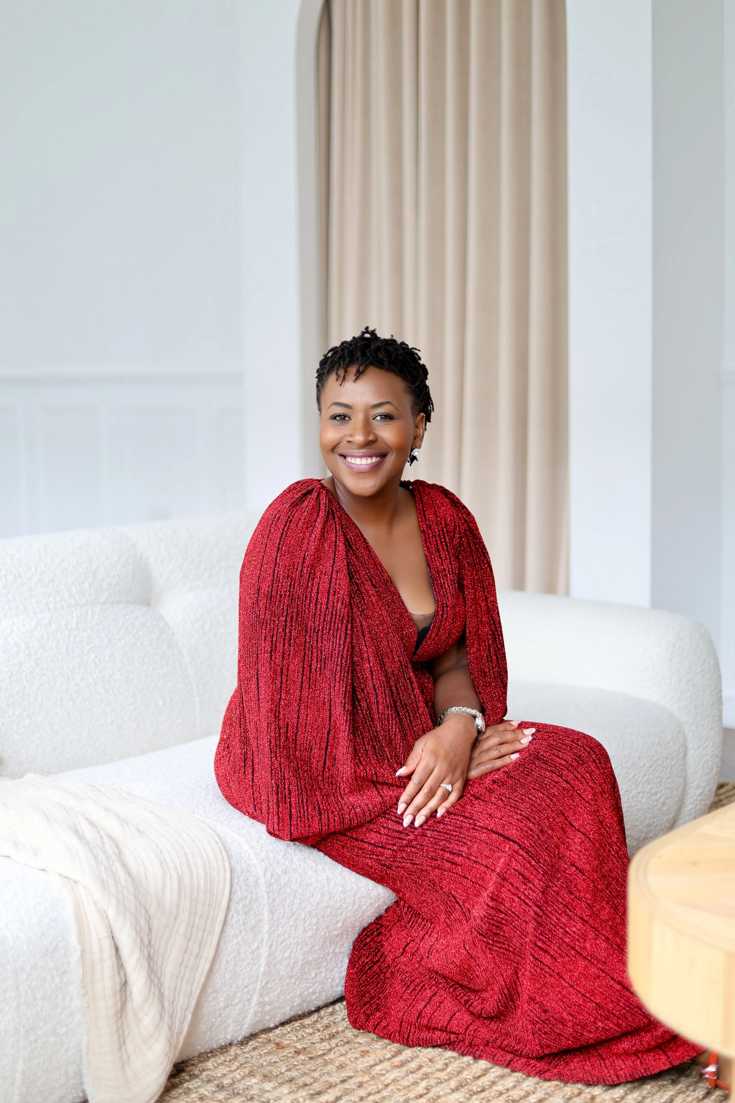 A woman in a red dress sitting on a white couch with beige curtains in the background, smiling at the camera.