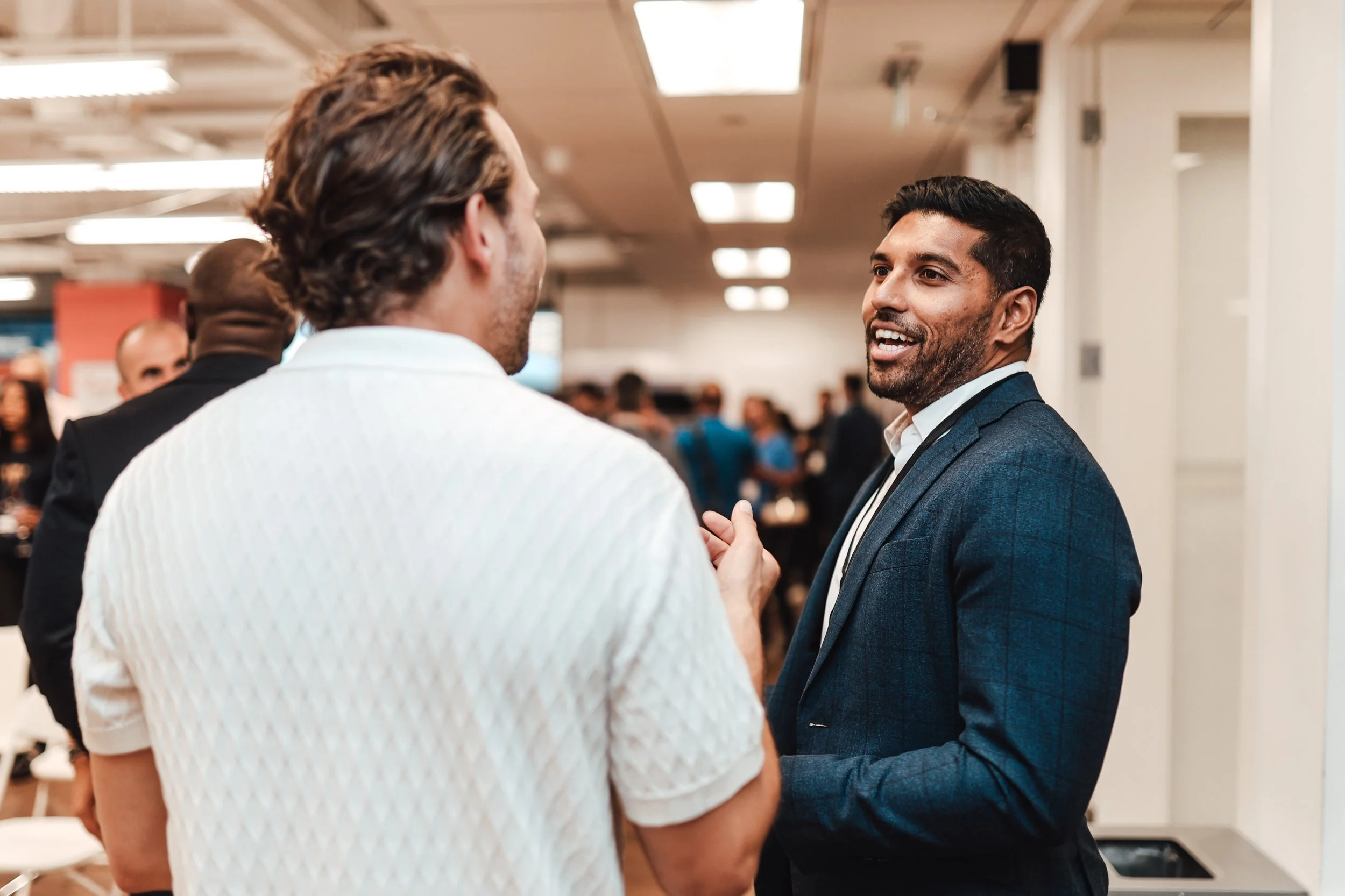 Two men are engaged in conversation at a networking event. One man, with darker hair, facial hair, and wearing a dark blue suit, is smiling while talking. The other man, with lighter hair and wearing a white textured shirt, is facing away from the ca