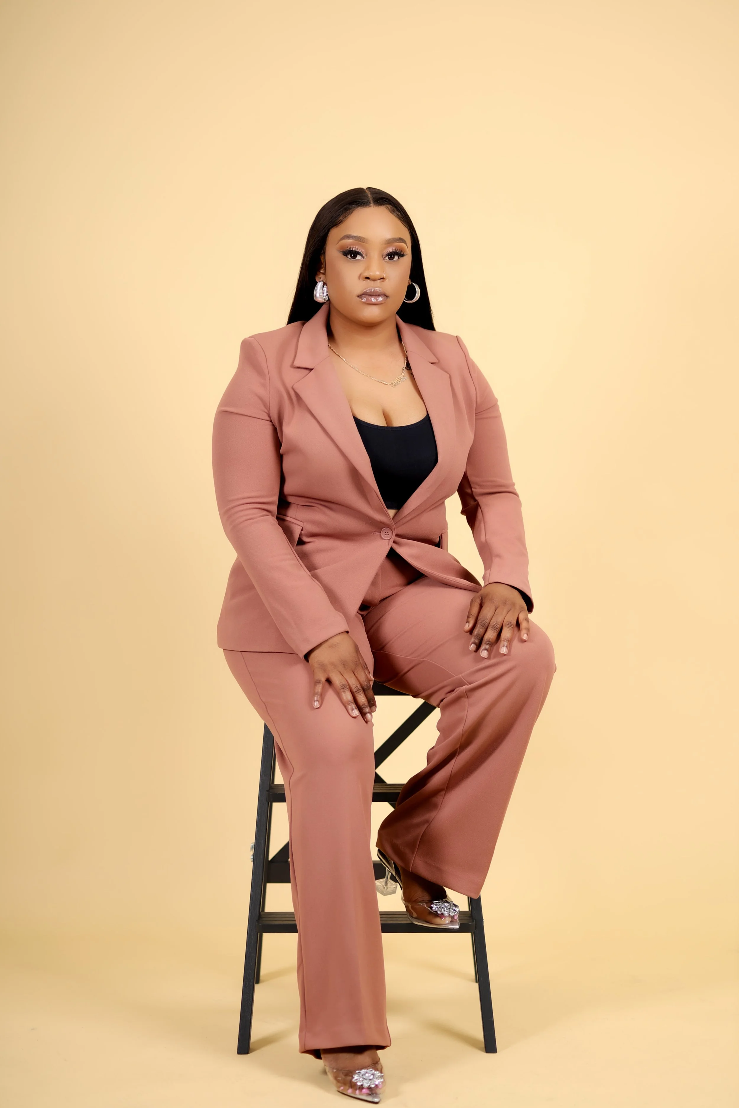 A woman sitting on a black stool against a beige background, wearing a pink suit, black top, and high heels.