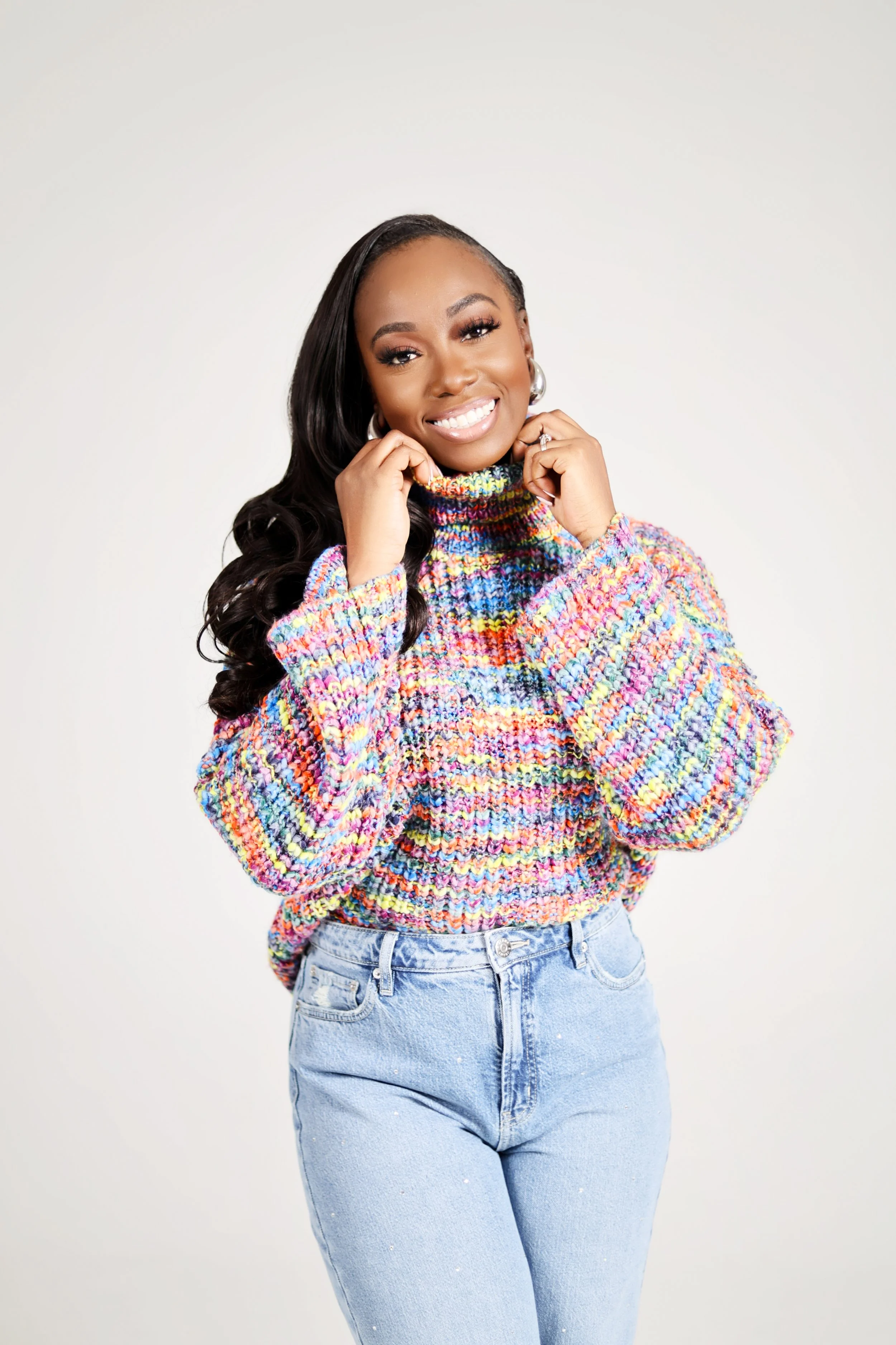 A woman with long black hair wearing a colorful knitted sweater and light blue jeans, smiling and posing with her hands near her face against a plain light grey background.