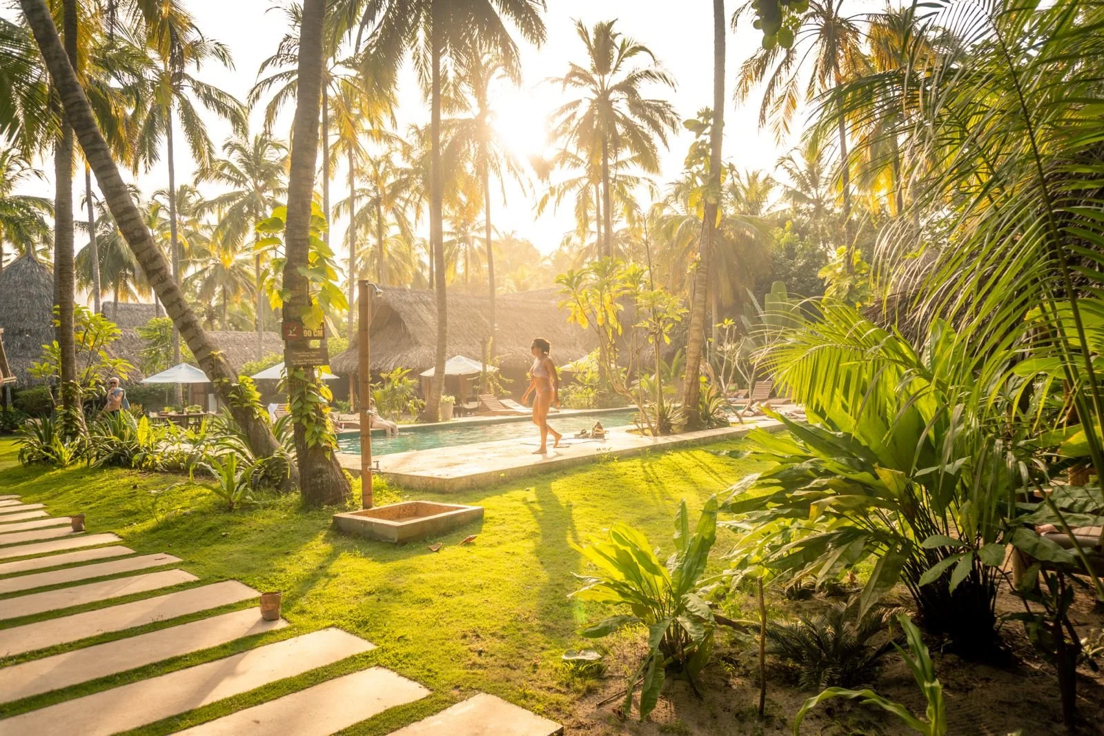Outdoor pool area with lounge chairs, palm trees, string lights, and thatched roof structures.