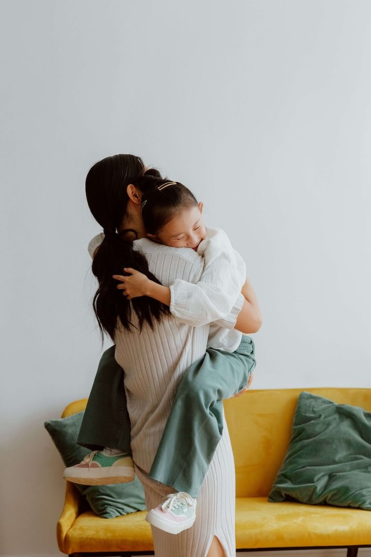Mother hugging her smiling young daughter in front of a mustard-yellow sofa representing connection after supportive therapy session.
