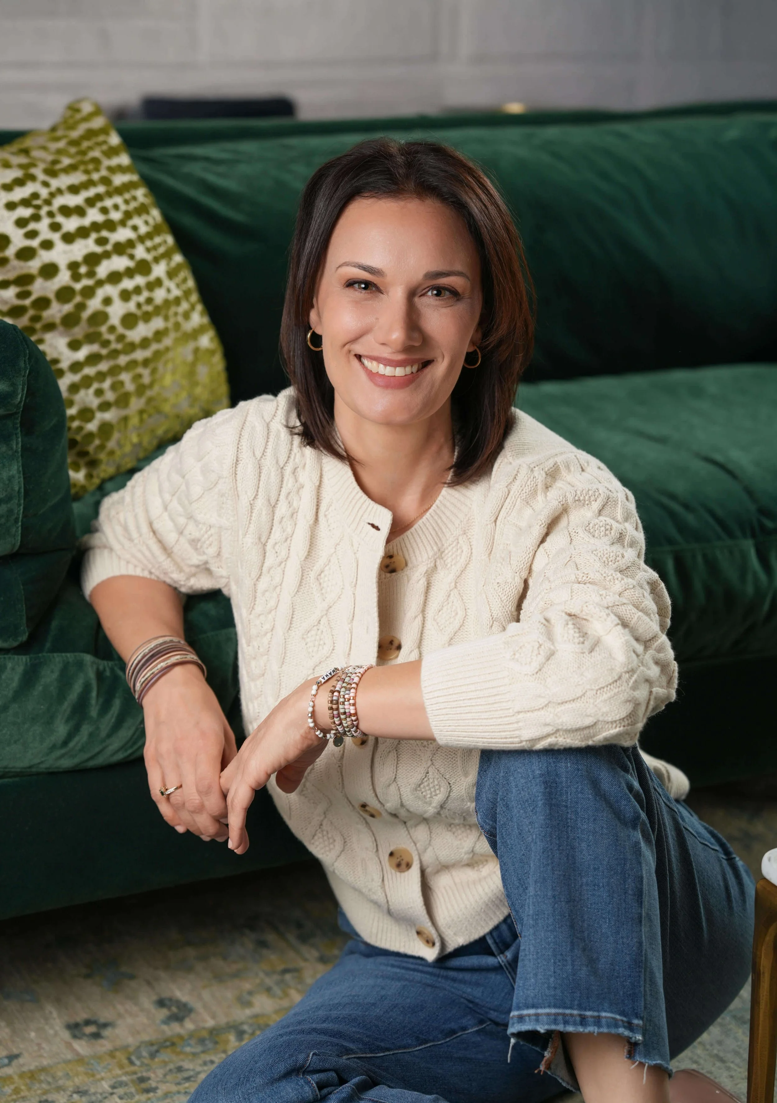 Sara Havlik, LMSW in a cream cable-knit cardigan sitting on a dark green couch