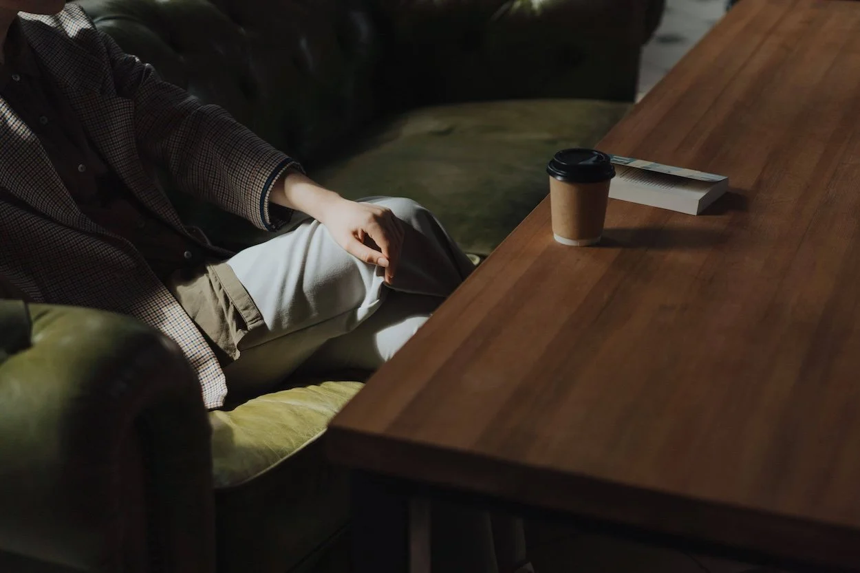 A person sitting in an armchair beside a wooden table with takeaway coffee and a book, as if in an adult therapy session.