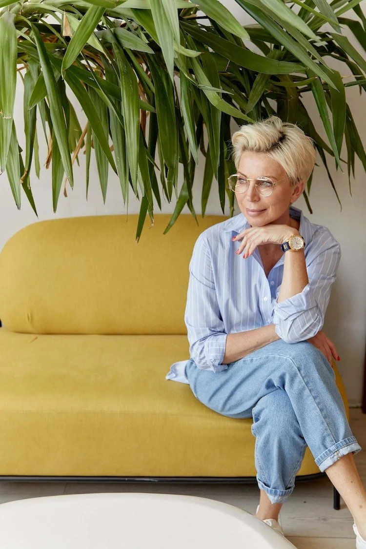 Stylish older woman with short blonde hair sitting on a mustard-yellow sofa beneath a large leafy houseplant.