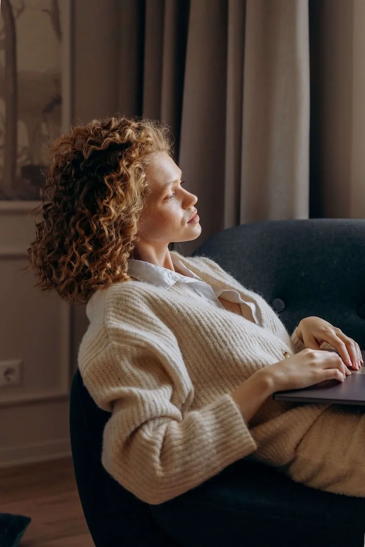 Woman with curly hair relaxing in an armchair by a window with a laptop after a virtual therapy session for OCD.