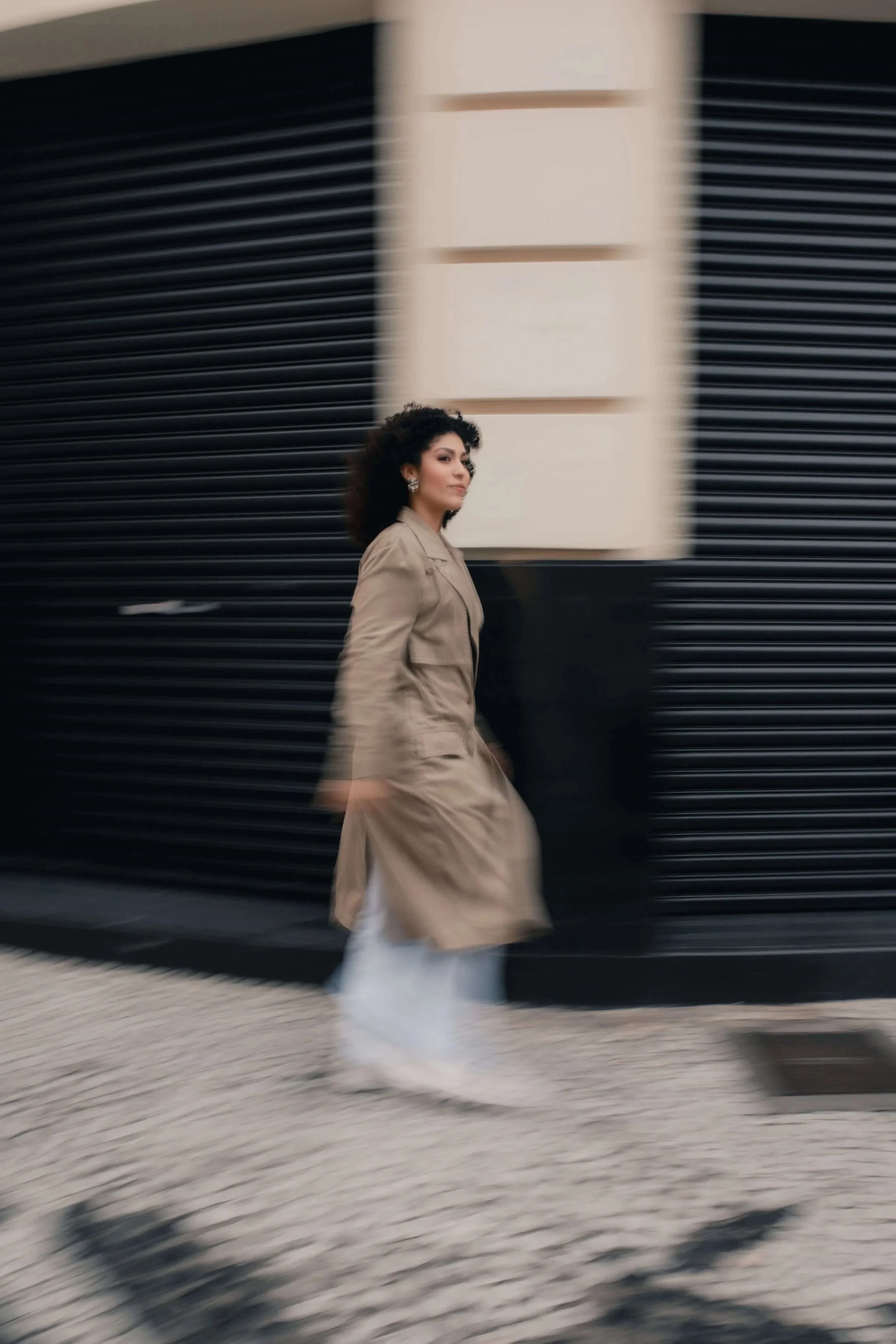 Woman with dark, curly hair in tan trench coat walking past a black textured wall on a street with a blurred camera perspective