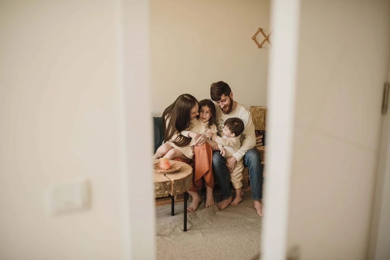 Family of four sitting together on a couch, photographed through a partially open doorway, representing adoption-competent therapy outcomes.