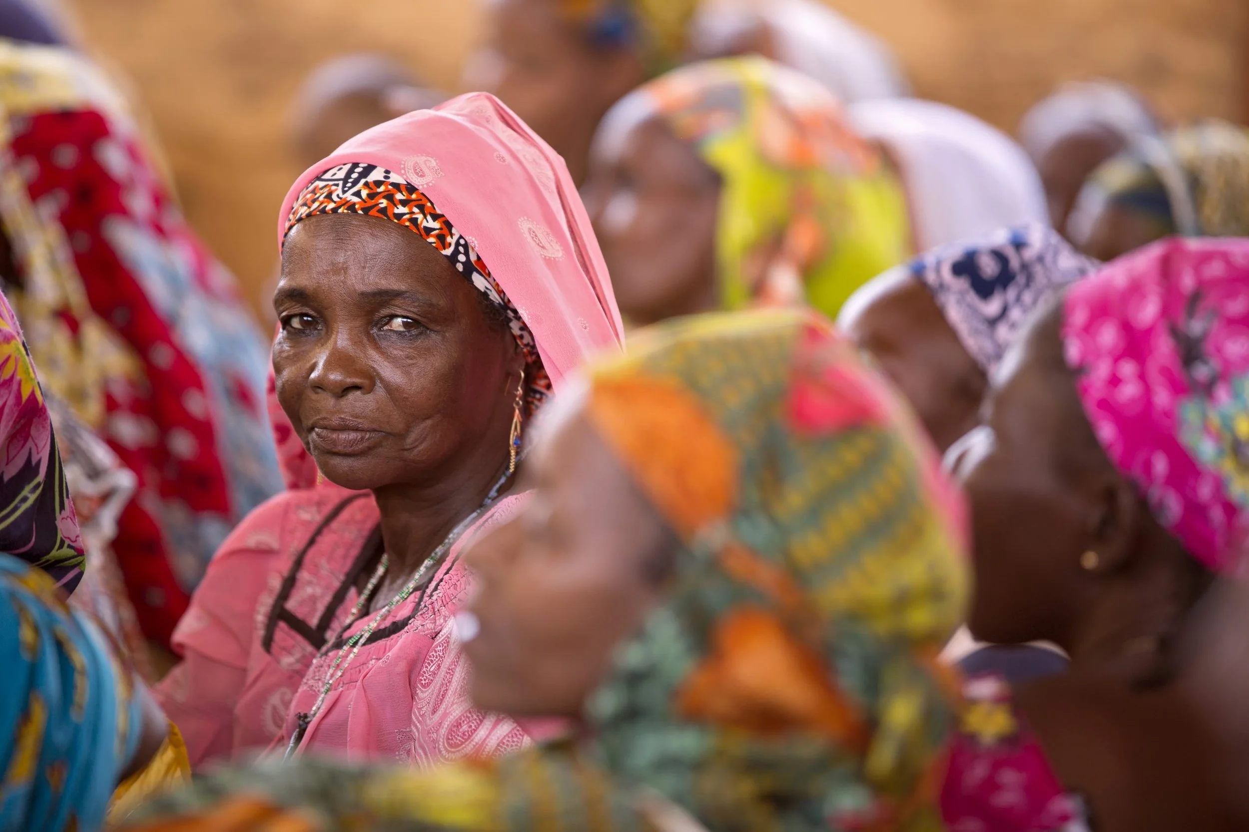 Group of women in colorful traditional attire, focus on a woman in pink hijab.