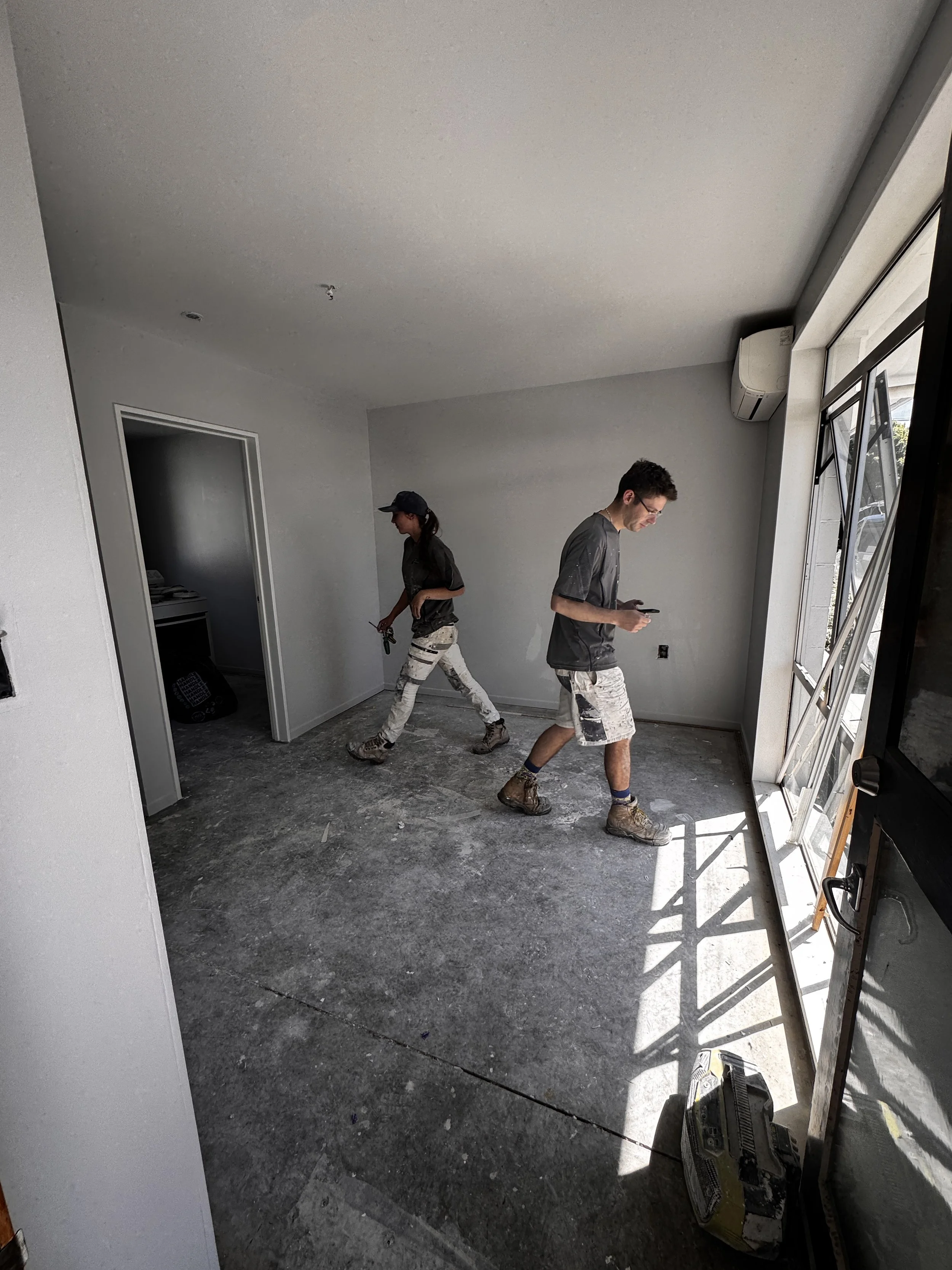 Two workers in construction attire walking inside a room with unfinished concrete floors, white walls, and a large window letting in natural light; construction equipment visible.