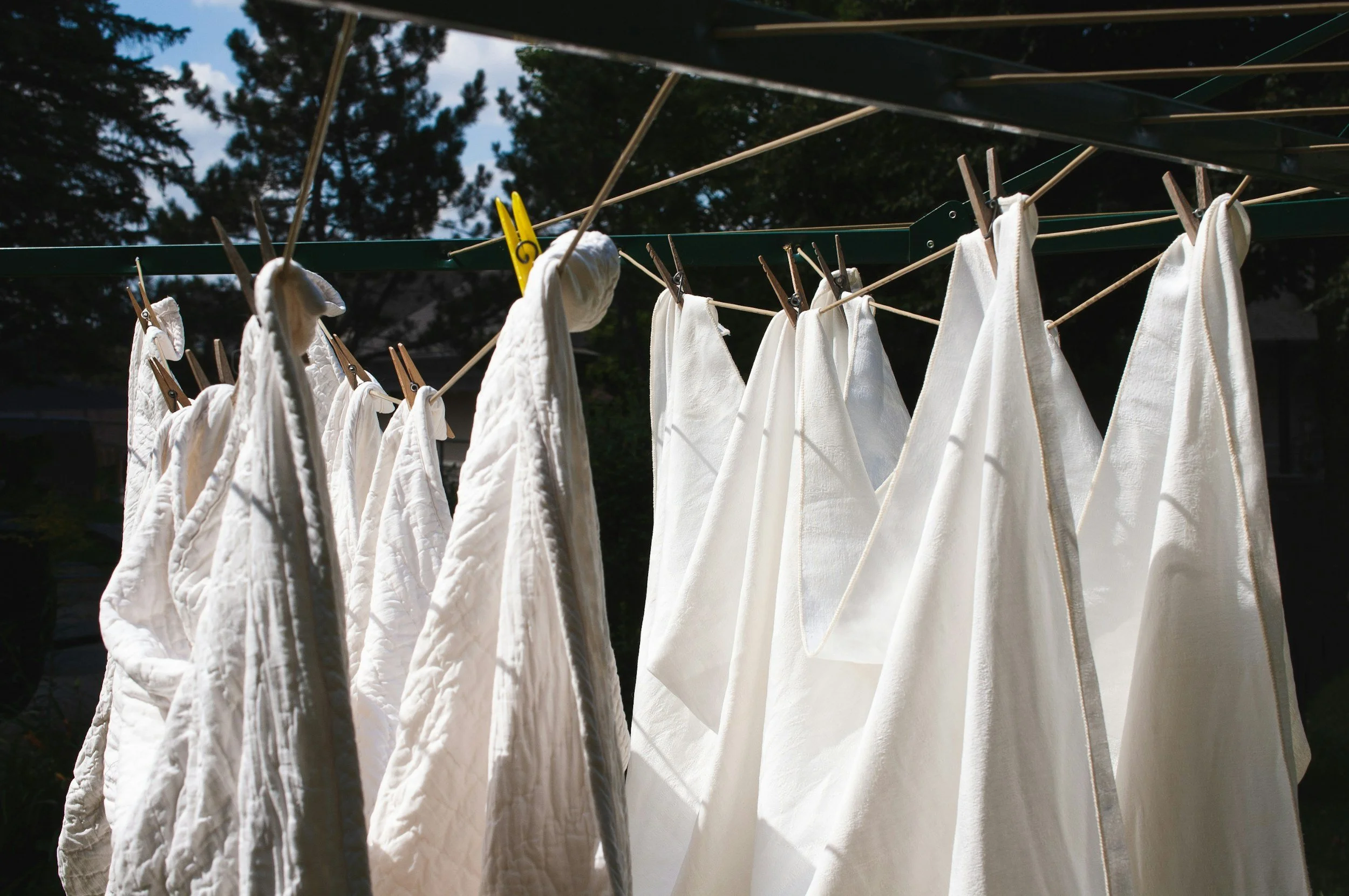 White laundry hanging on a clothesline with wooden clothespins outside, with trees and a cloudy sky in the background.
