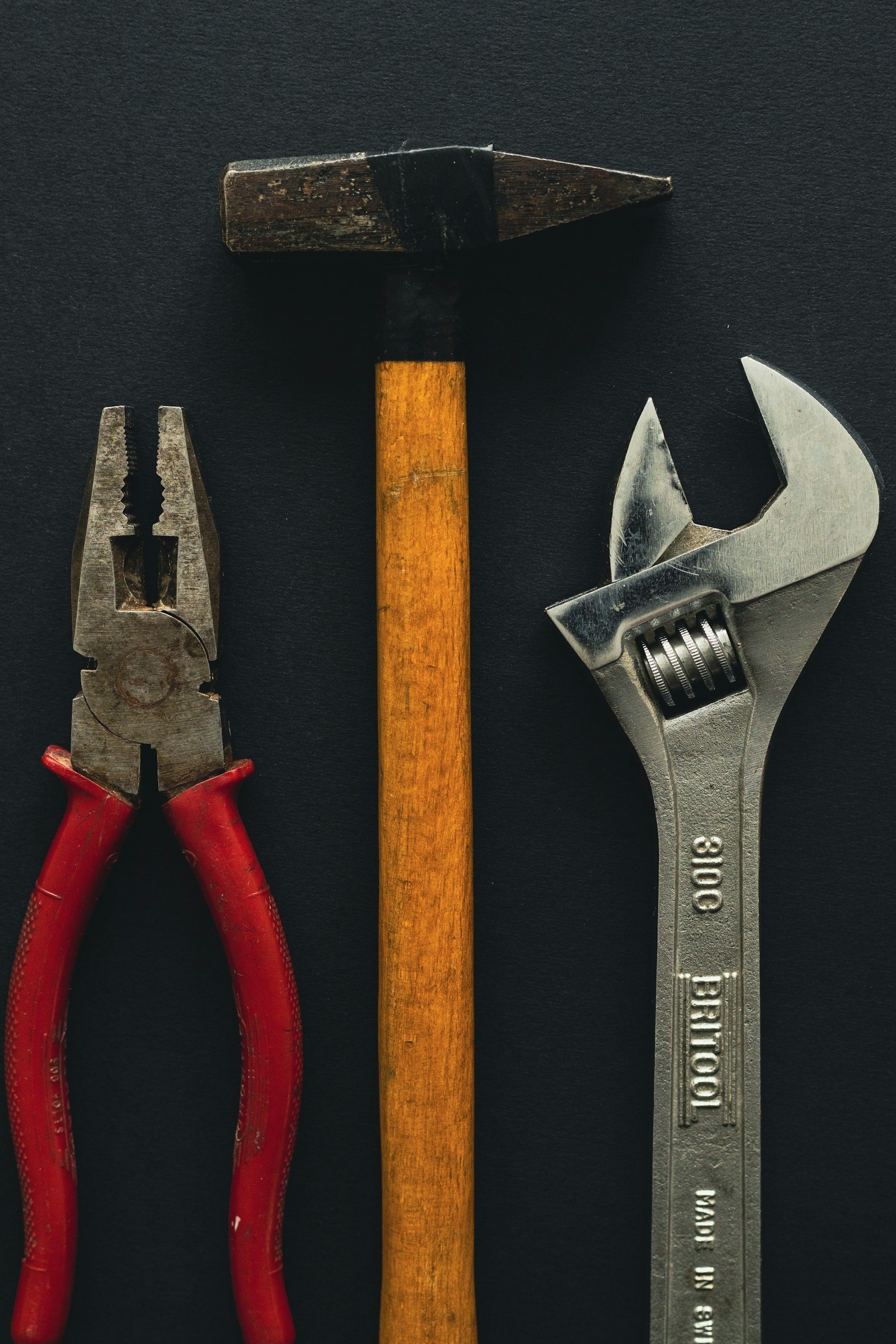 A set of three tools arranged vertically: channel lock pliers with red handles on the left, a hammer with a wooden handle in the middle, and an adjustable wrench on the right, all on a black background.