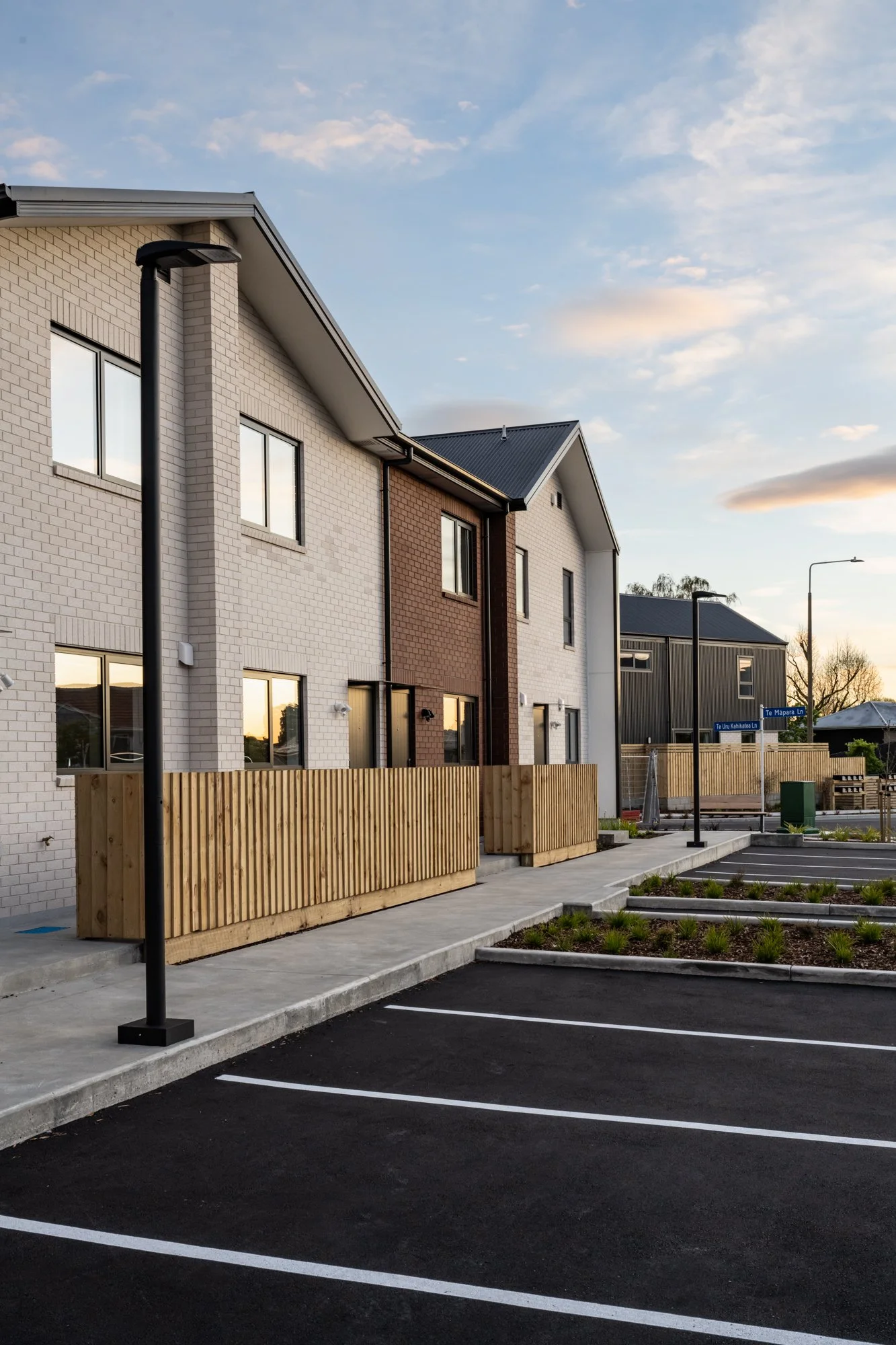 A row of modern townhouses with white and brown brick exteriors, black roofs, wooden fences, and a parking lot in the foreground on a clear evening.