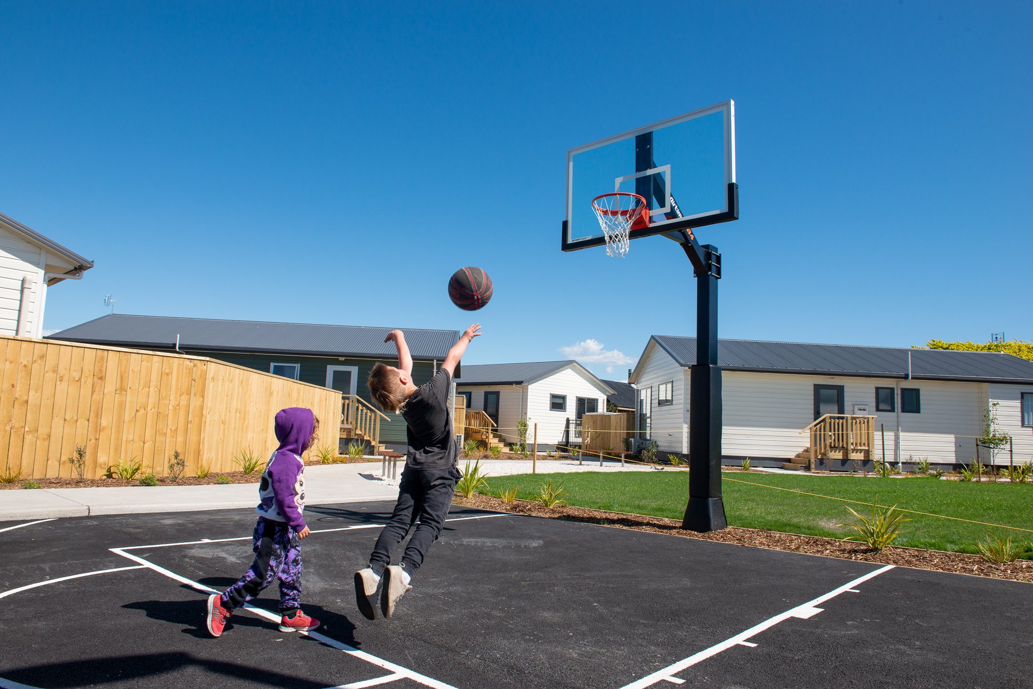Two children playing basketball outdoors on a sunny day, one jumping to shoot or block the ball near the hoop, with houses and a wooden fence in the background.