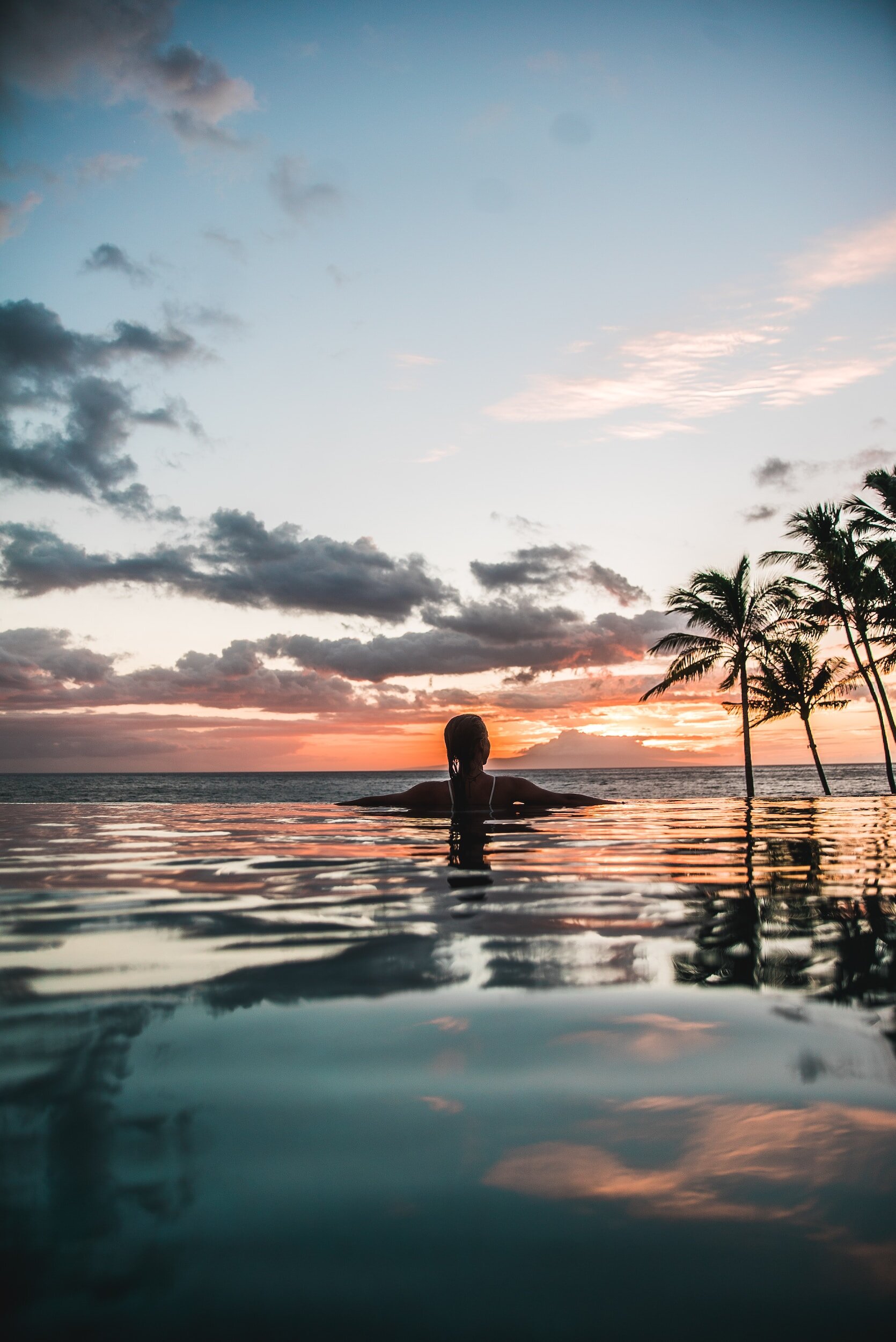 A person swimming in an infinity pool at sunset with palm trees and a cloudy sky in the background.