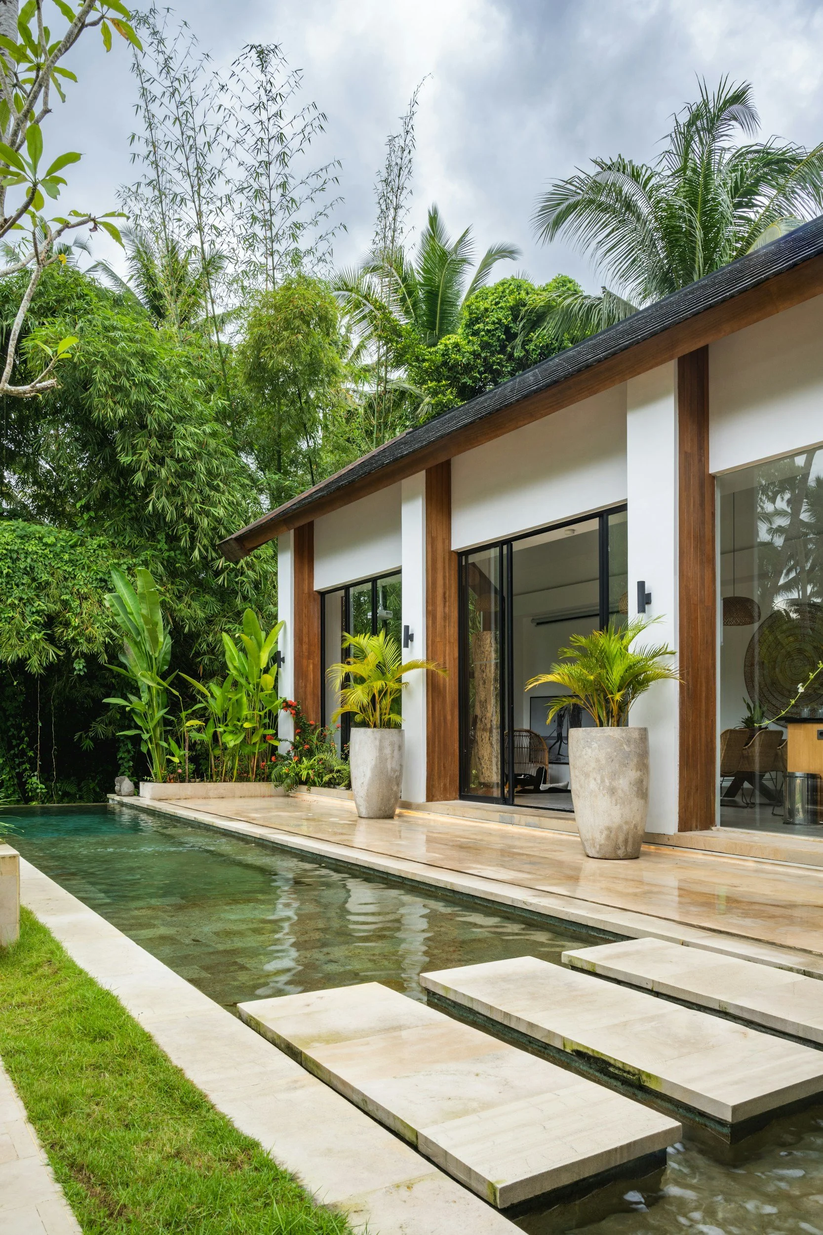 Modern house with large glass doors, potted plants, and a narrow water feature with stepping stones, surrounded by lush green tropical trees and plants.