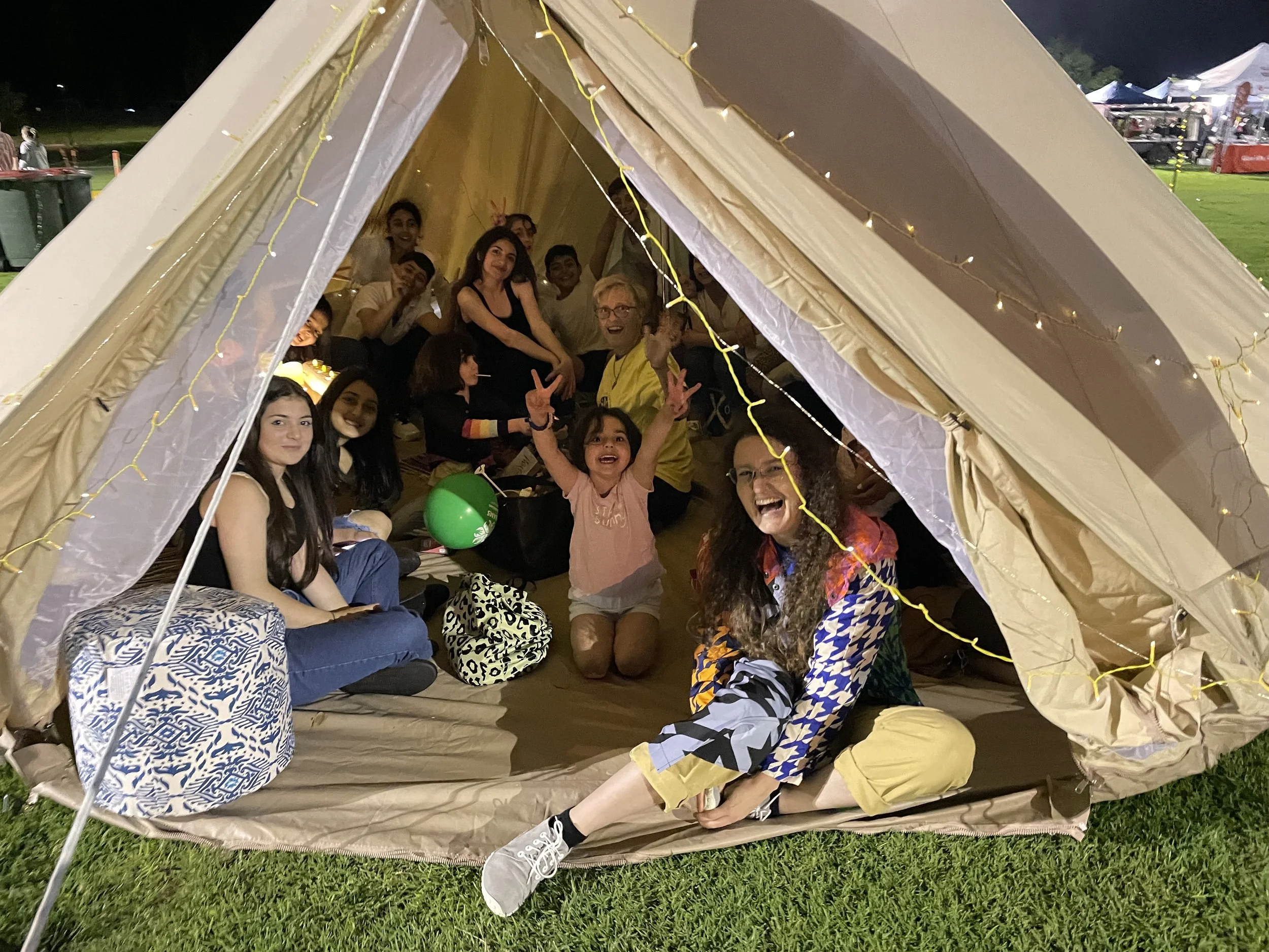 A group of children and adults inside a lit tent at night, smiling and making peace signs, with some sitting on the ground and others on benches, and a grassy area outside.