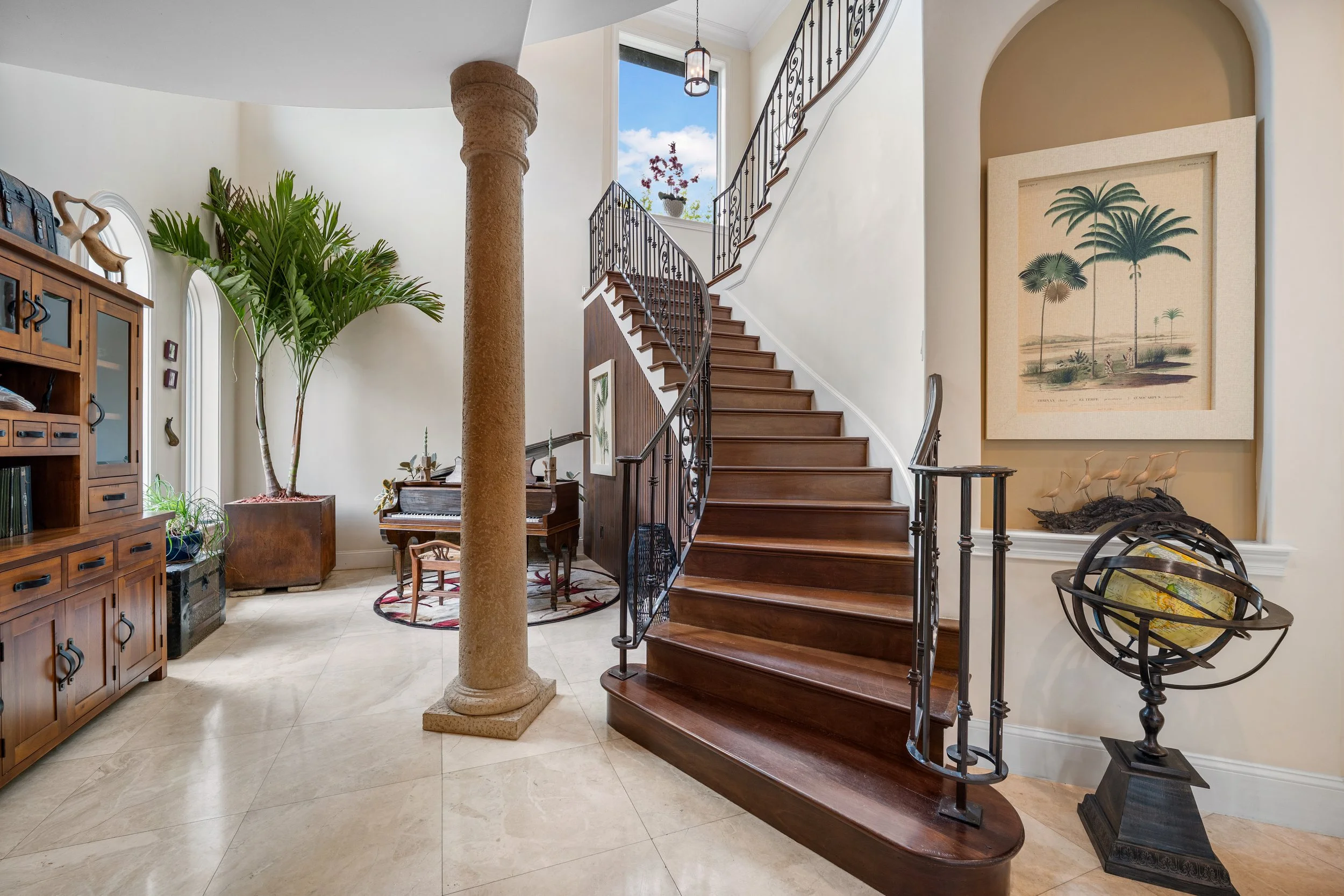 Elegant foyer featuring a curved wooden staircase, decorative columns, and warm travertine flooring.