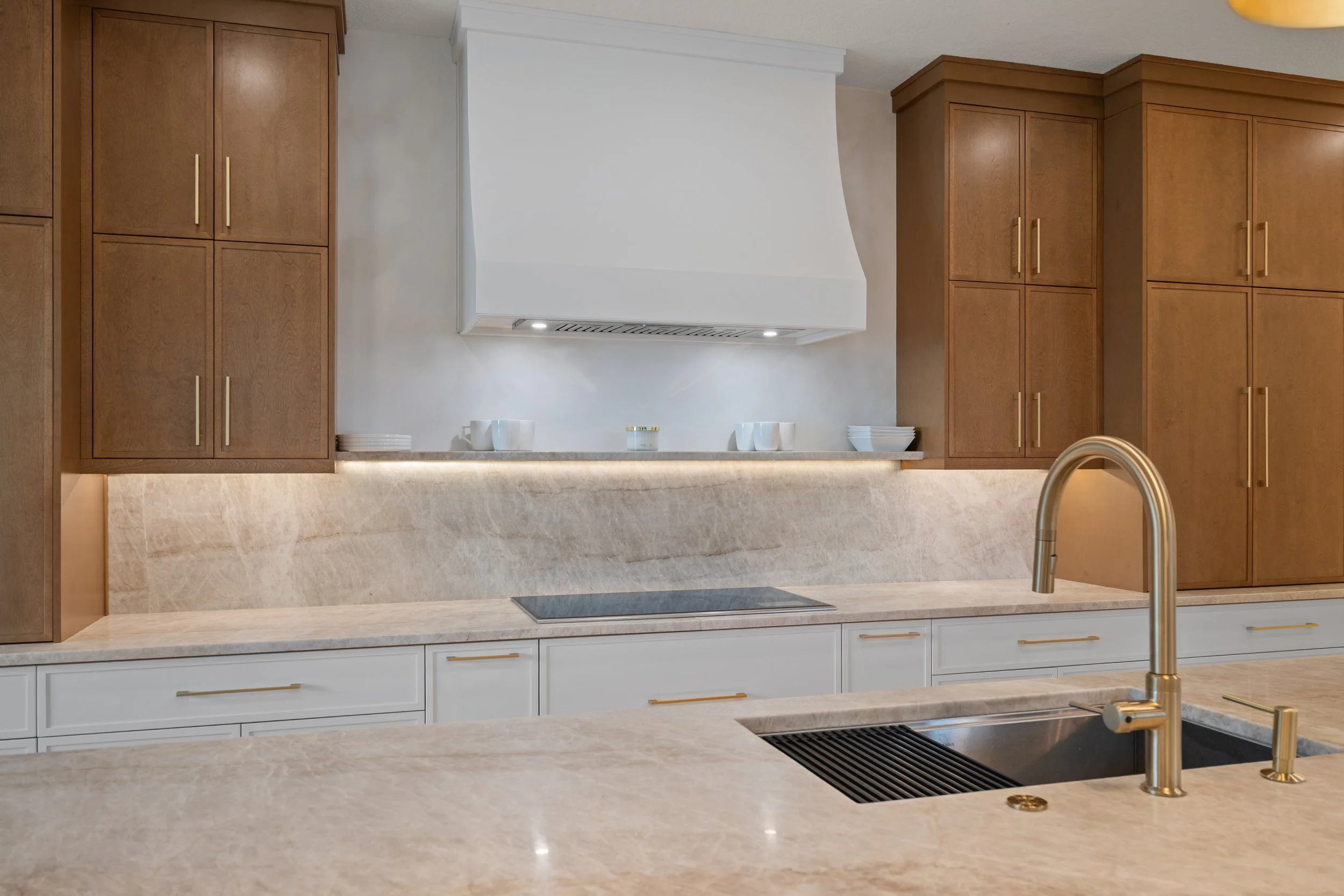 Modern kitchen with beige marble countertops and backsplash, wooden cabinets, and a built-in stainless steel oven and cooktop. A brass faucet is visible at the foreground sink.