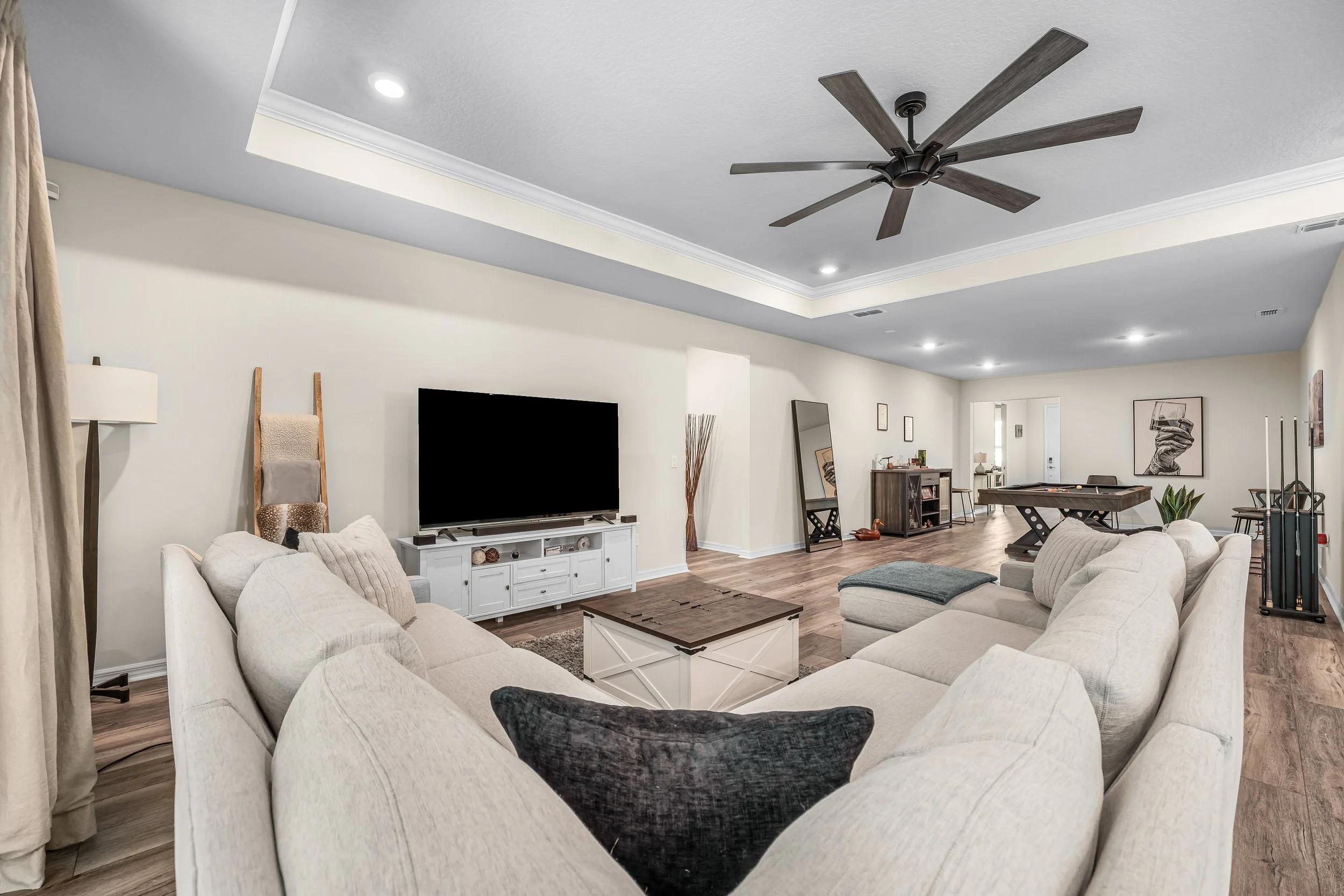 A spacious living room with beige sofas arranged in a U-shape, a white TV stand with a large flat-screen TV, a coffee table, a pool table, and various artwork and decor items.