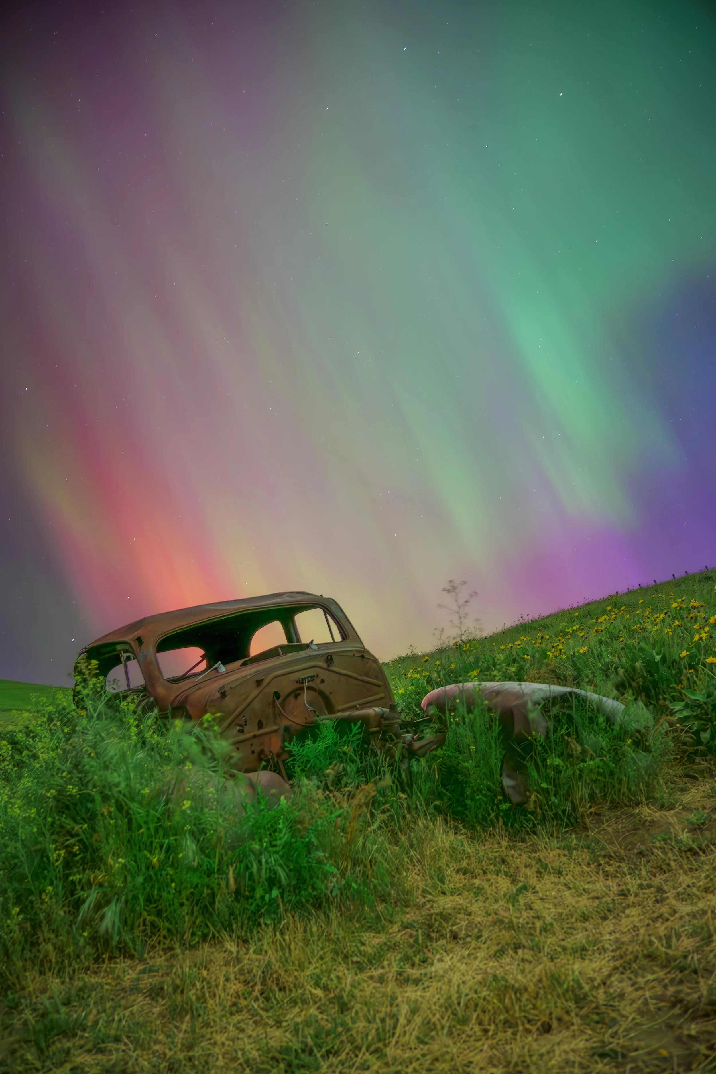Rusty car wreckage in a grassy field under a colorful night sky with aurora borealis.