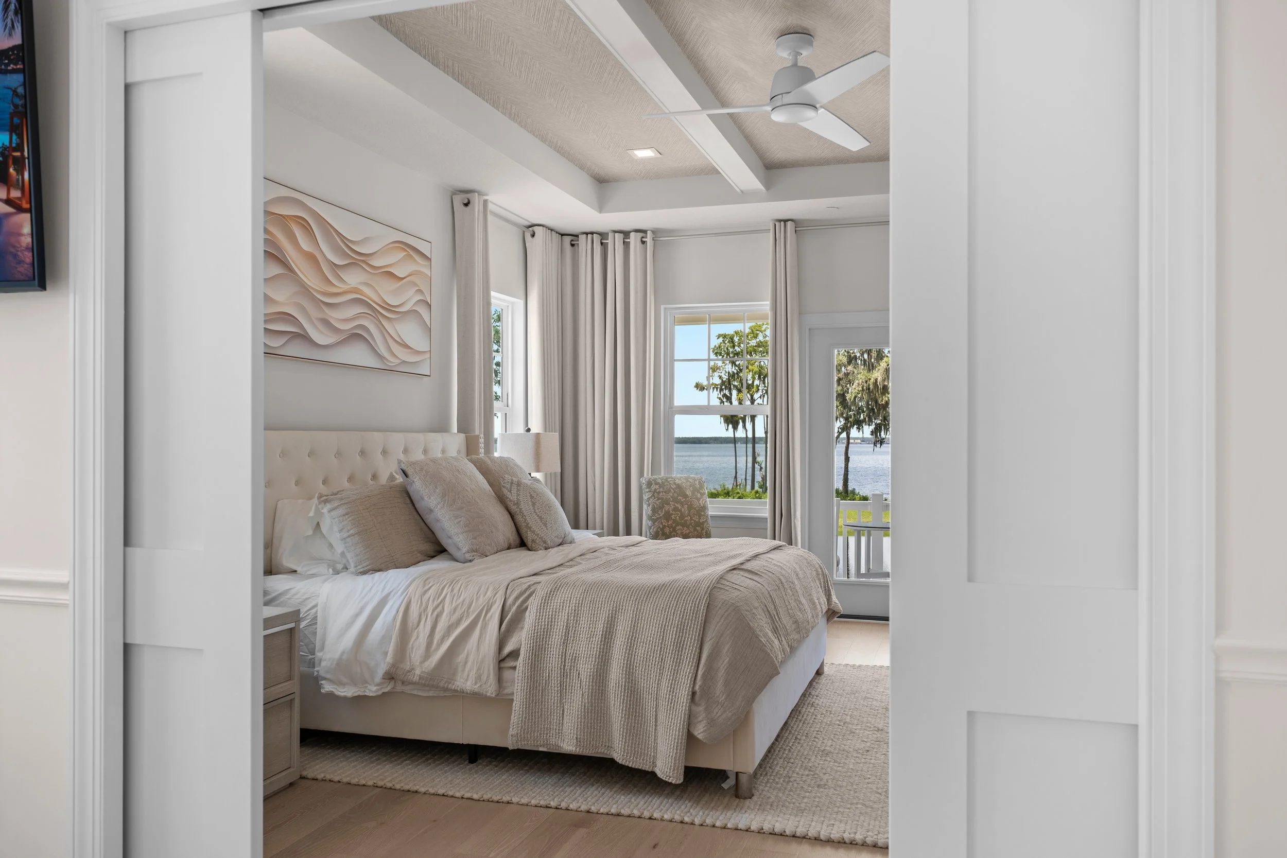 A bedroom viewed through a white, open doorway. The bed has a beige headboard and is made with beige and white bedding. There are light-colored pillows on the bed. Windows with white curtains show an outdoor scene with trees and water. A wall art pie