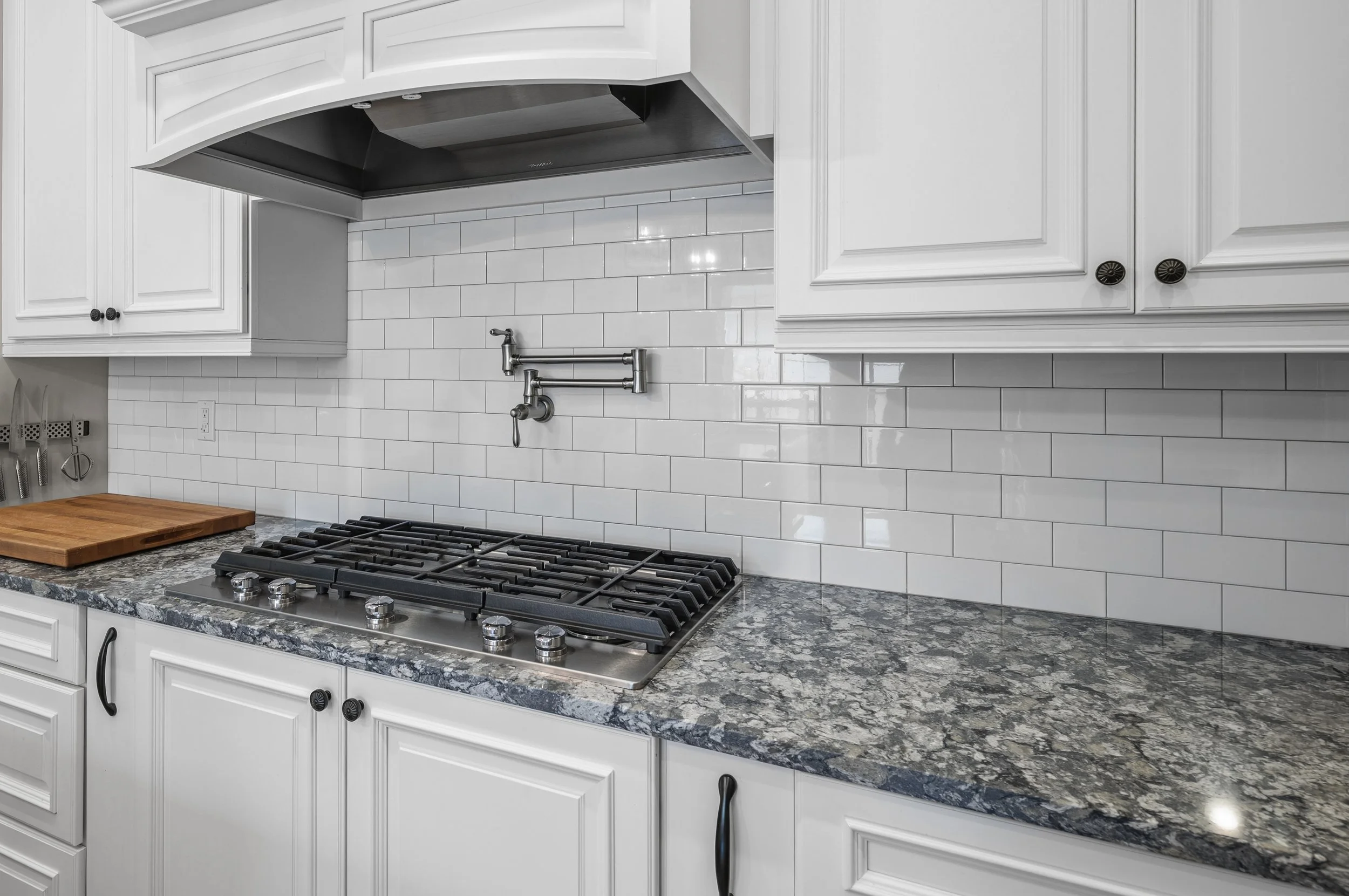 White kitchen cabinets with black knobs, a gas stove with four burners, a granite countertop, a cutting board, a white subway tile backsplash, and a black range hood.