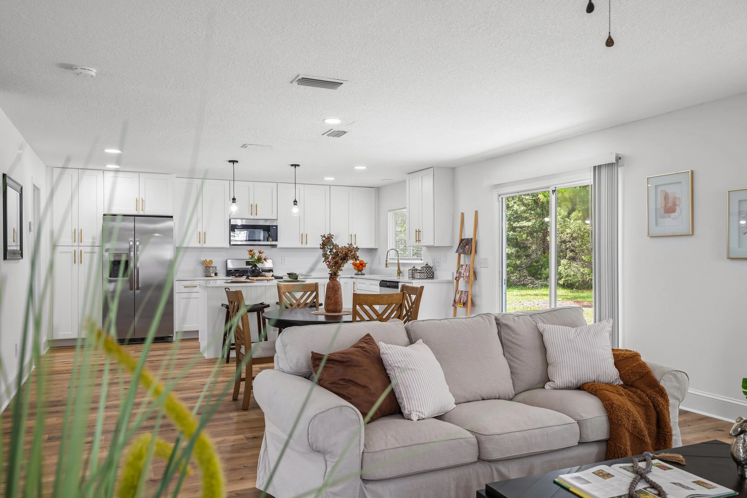 Open-concept living area with a white kitchen, wood floors, and a cozy sectional facing large windows to the backyard.