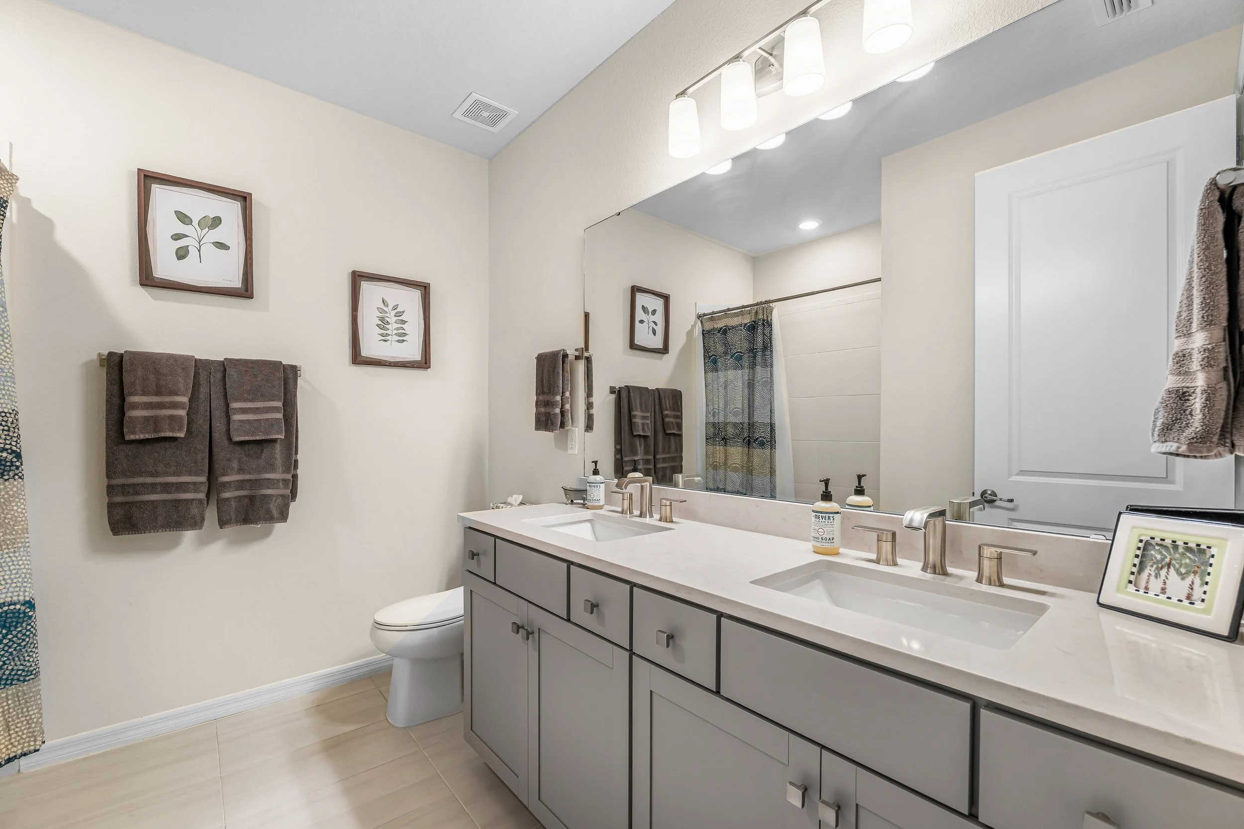 A modern bathroom with a large mirror, white countertops, and gray cabinets. There are two sinks, a soap dispenser, framed pictures, and three light fixtures above the mirror. Towels are hanging on the wall, and a shower with a patterned curtain is v