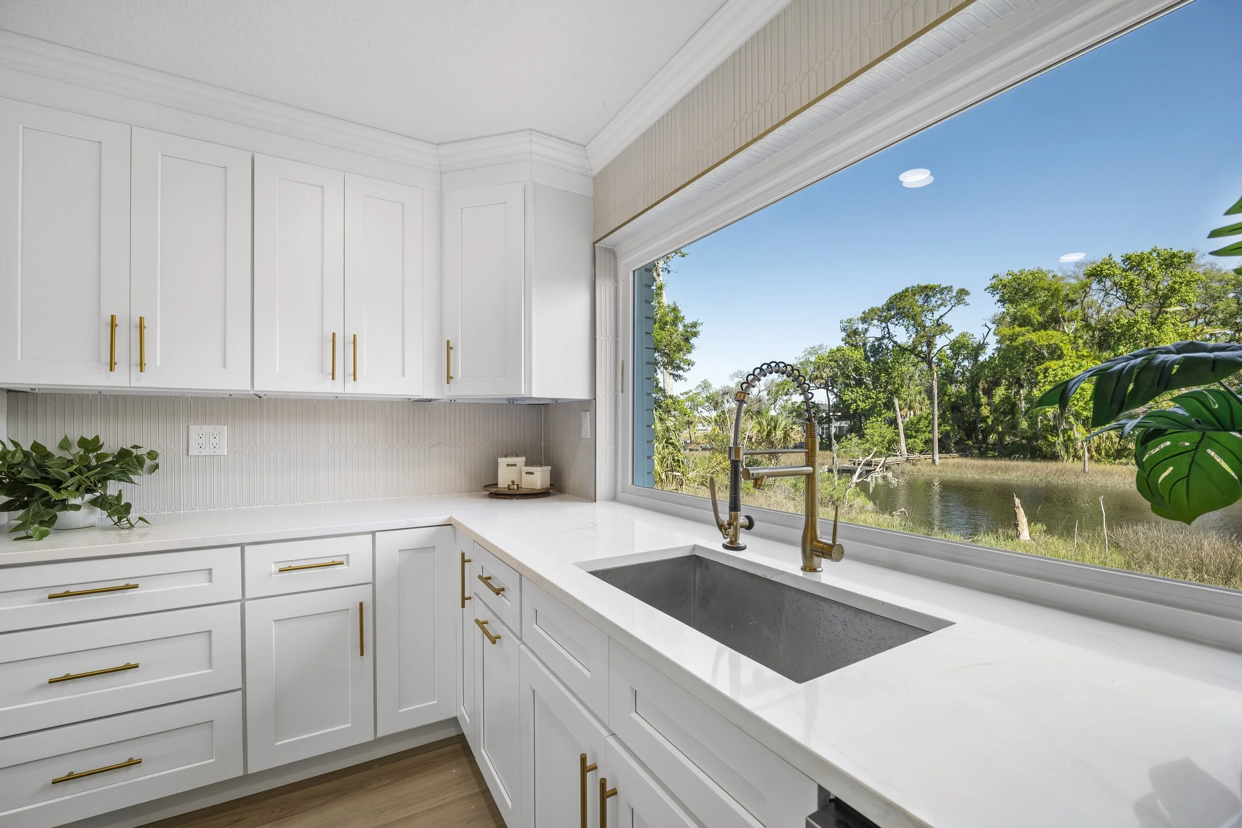 Bright white kitchen with a large window framing a wooded, waterfront view above the sink.