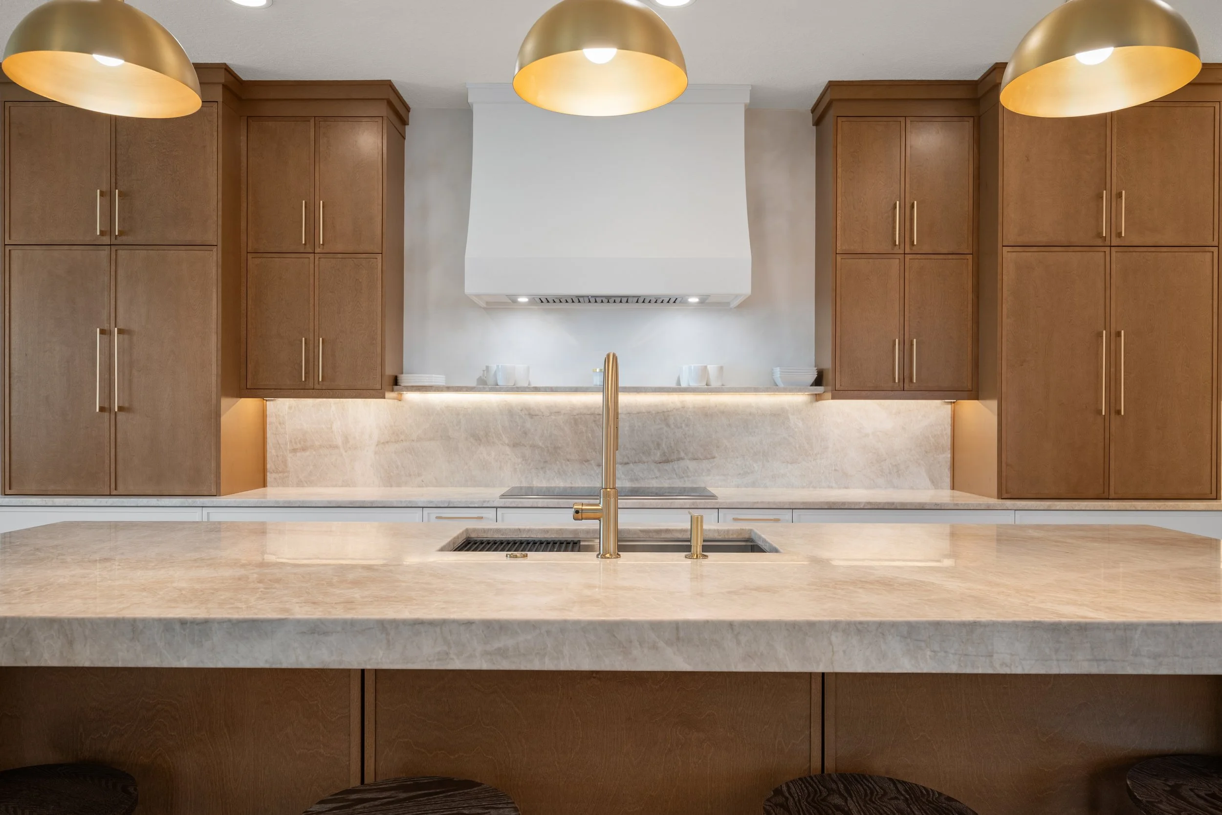 Kitchen with beige marble countertops, rich wood cabinetry, a gold pendant light, a white range hood, and a gold faucet.