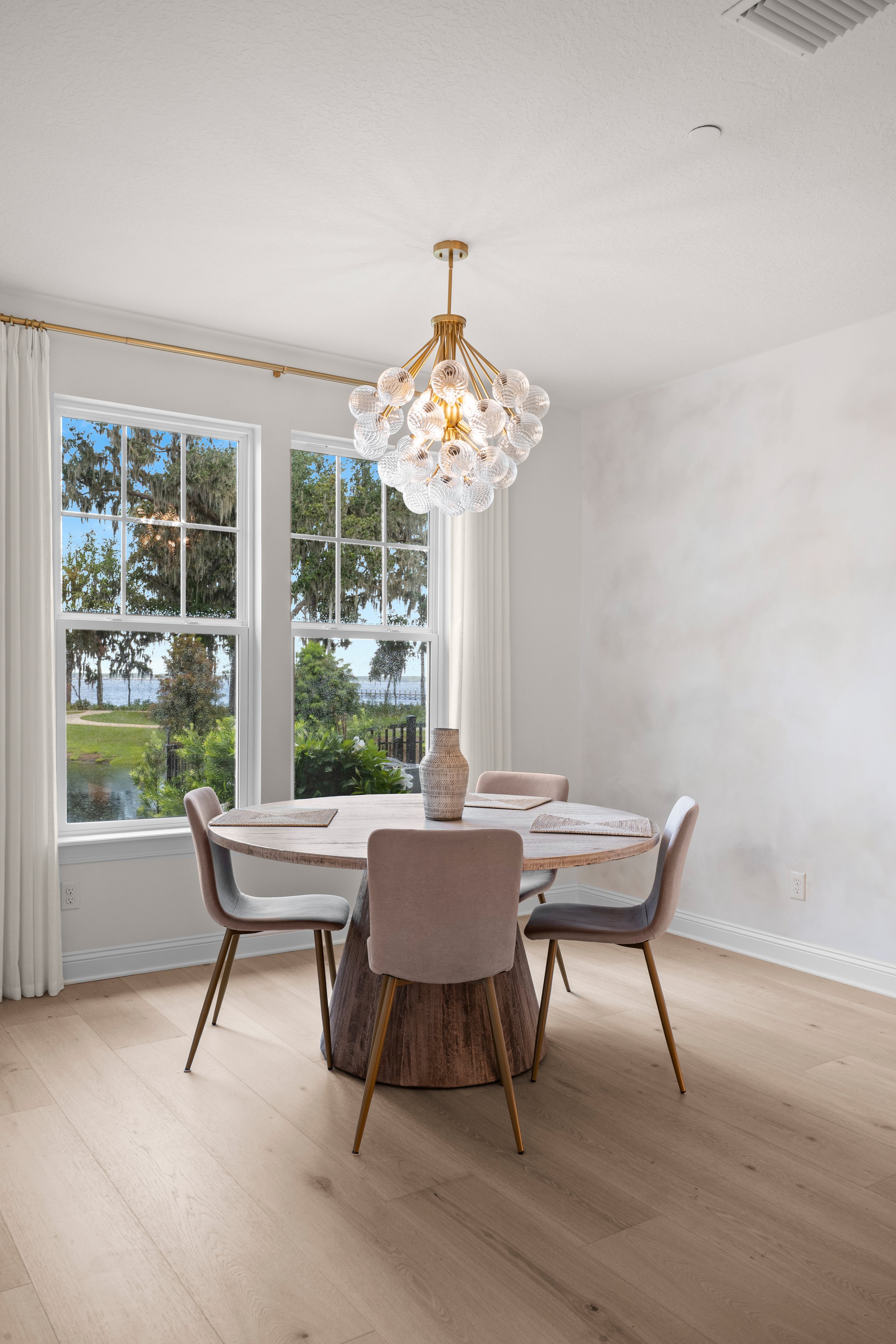 Dining room with round wooden table, six upholstered chairs, large window with curtains, chandelier with glass globes, and a view of greenery and water outdoor.