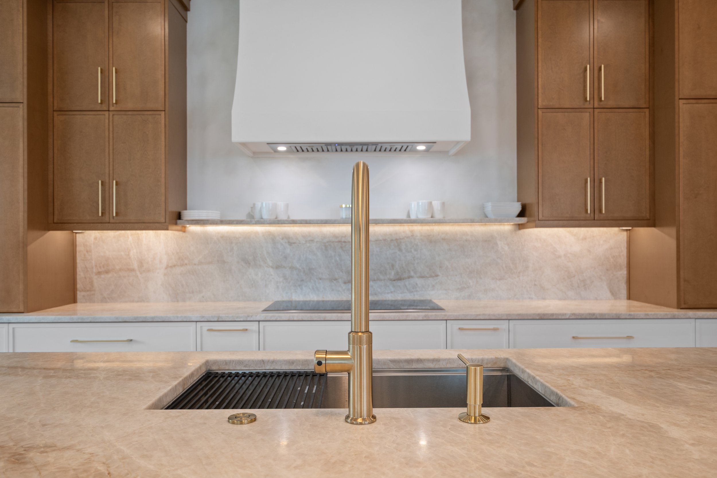 Modern kitchen with beige marble countertops, wooden cabinets with gold handles, a gold faucet, a white range hood, and white lower cabinets.