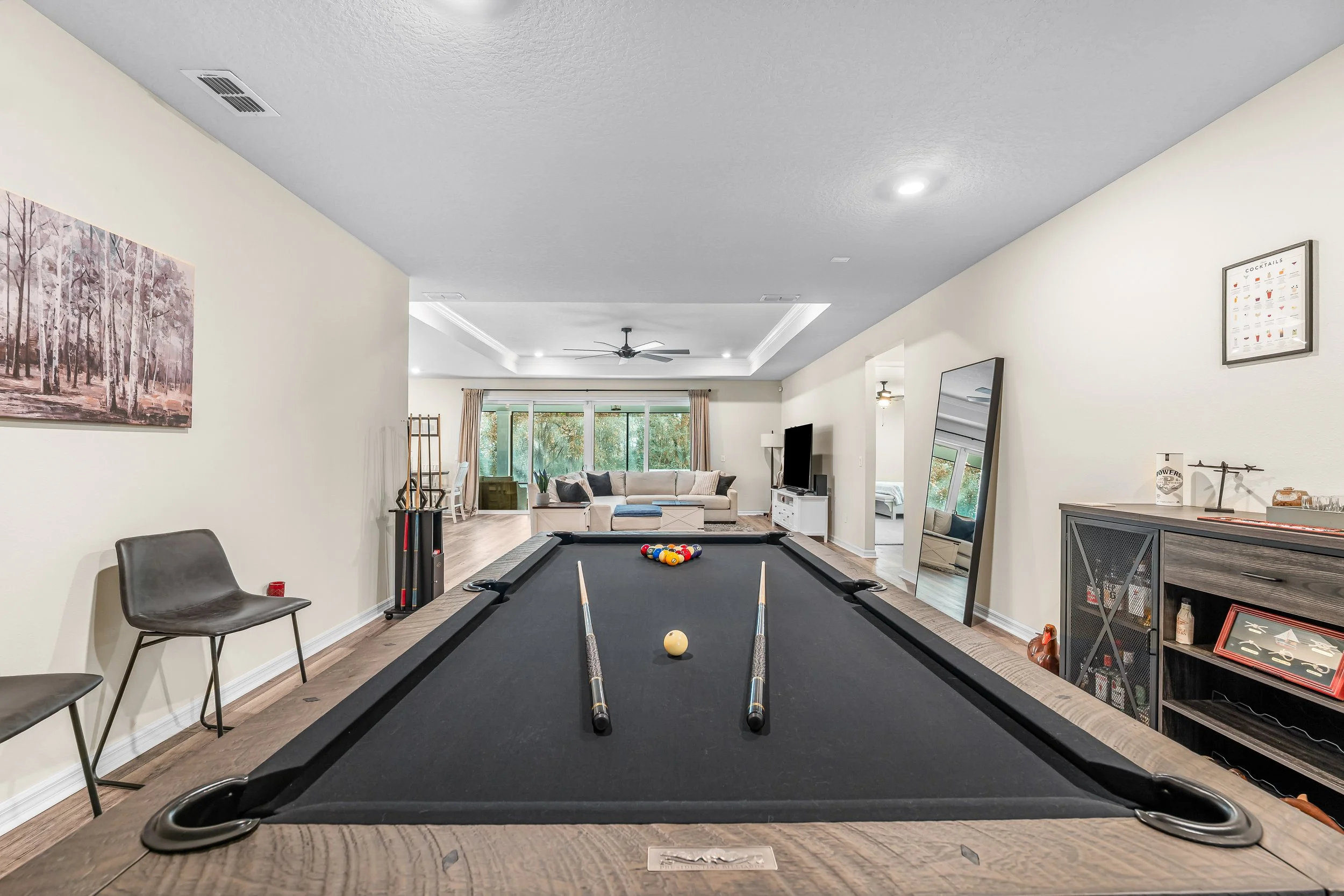 Interior of a living room with a pool table in the foreground, a large window with curtains, a beige sectional sofa, a ceiling fan, and various decor items including a mirror and wall art.