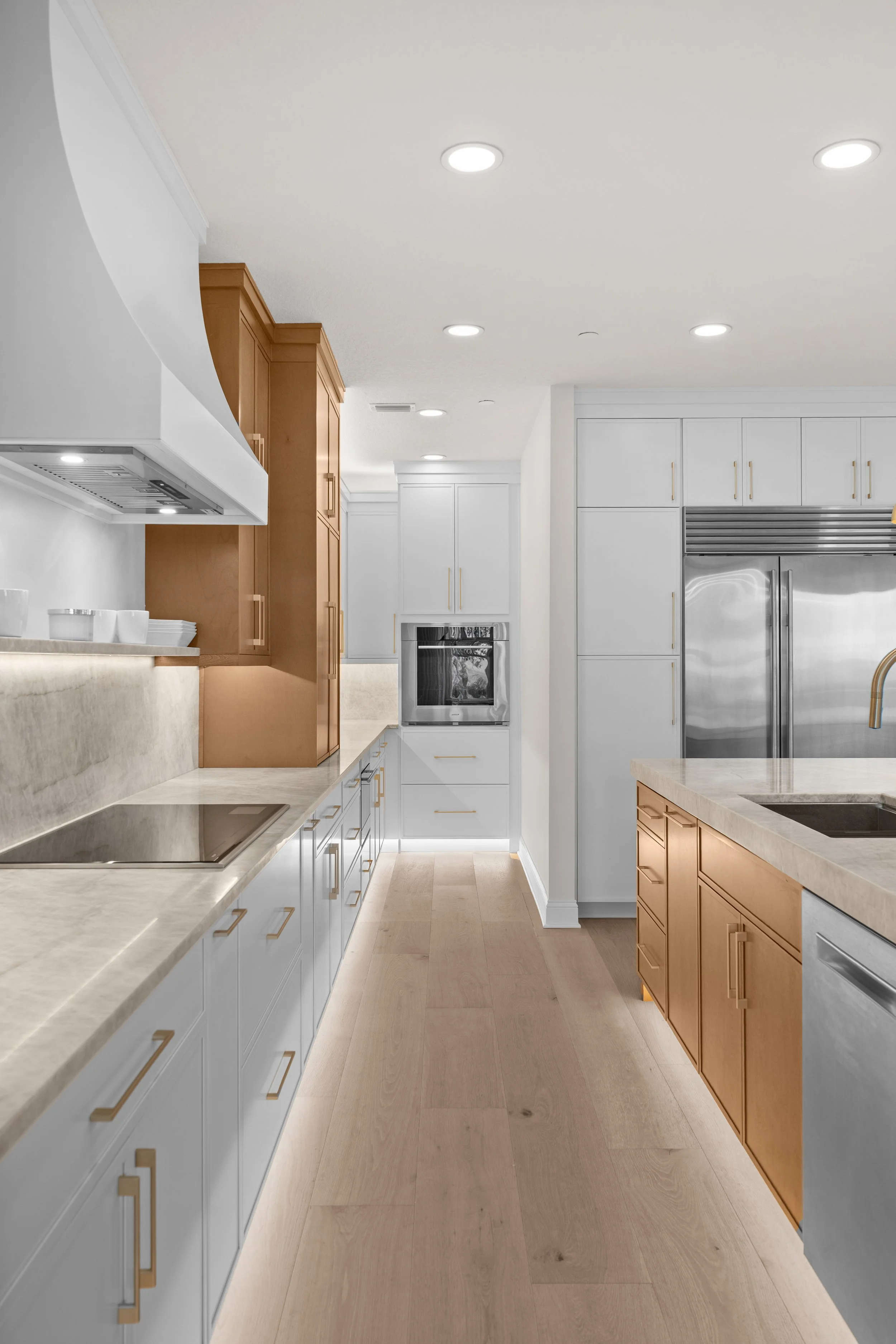 Modern kitchen with white and wooden cabinets, marble countertops, built-in oven, and stainless steel refrigerator, with hardwood floors and several recessed ceiling lights.