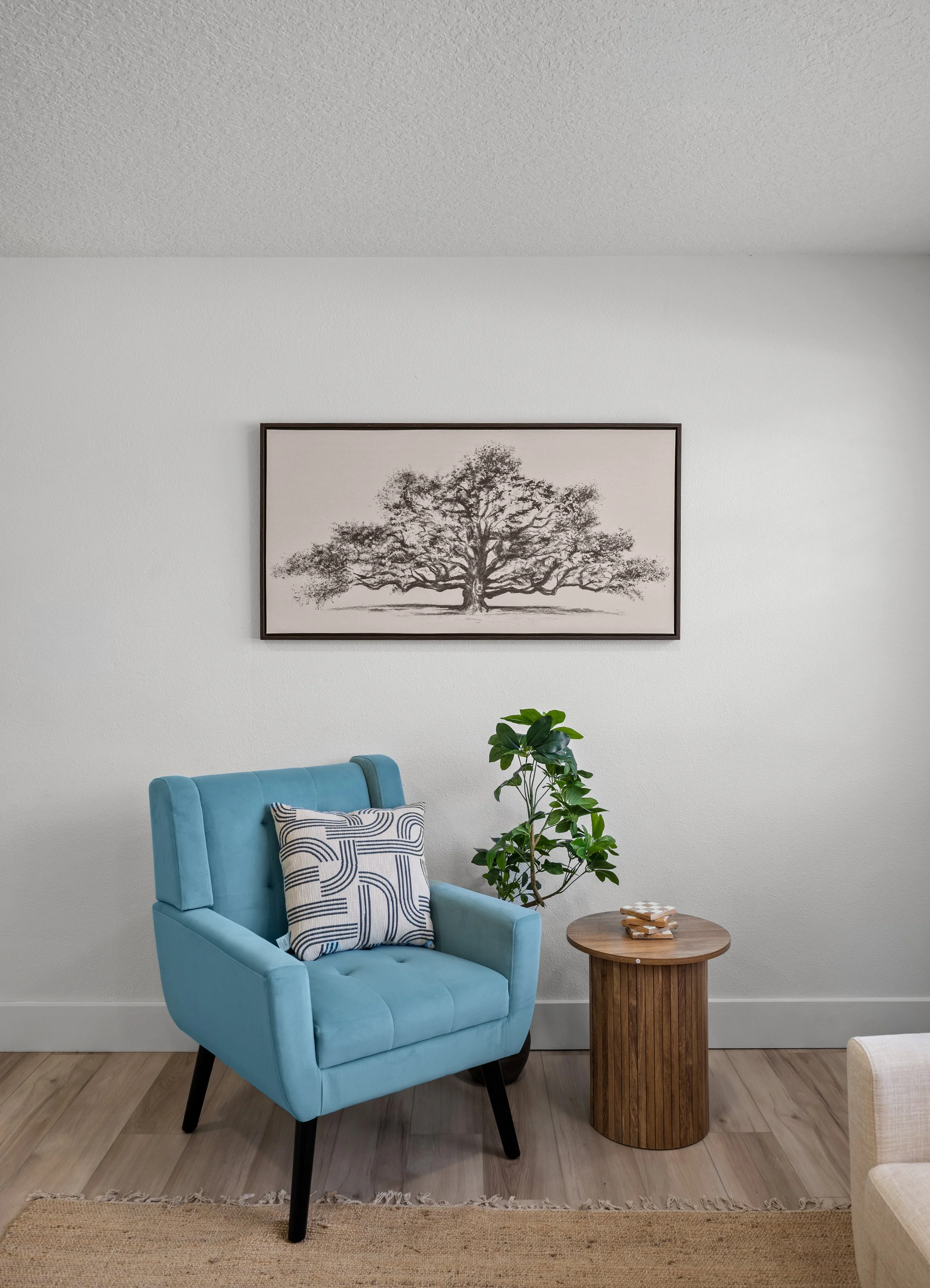 Interior of a living room with a light blue armchair, a patterned pillow, a wooden side table with coasters, a green plant, a framed black and white tree print on the wall, and a beige rug on a wooden floor.